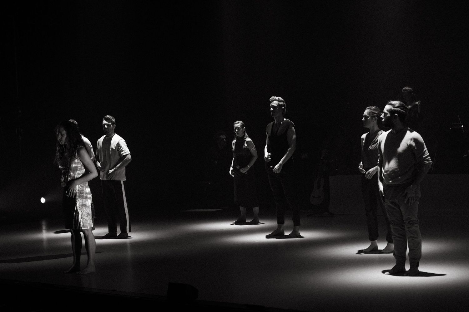 Several performers stand in dramatic spotlighting on a dark stage in black and white theatrical lighting.