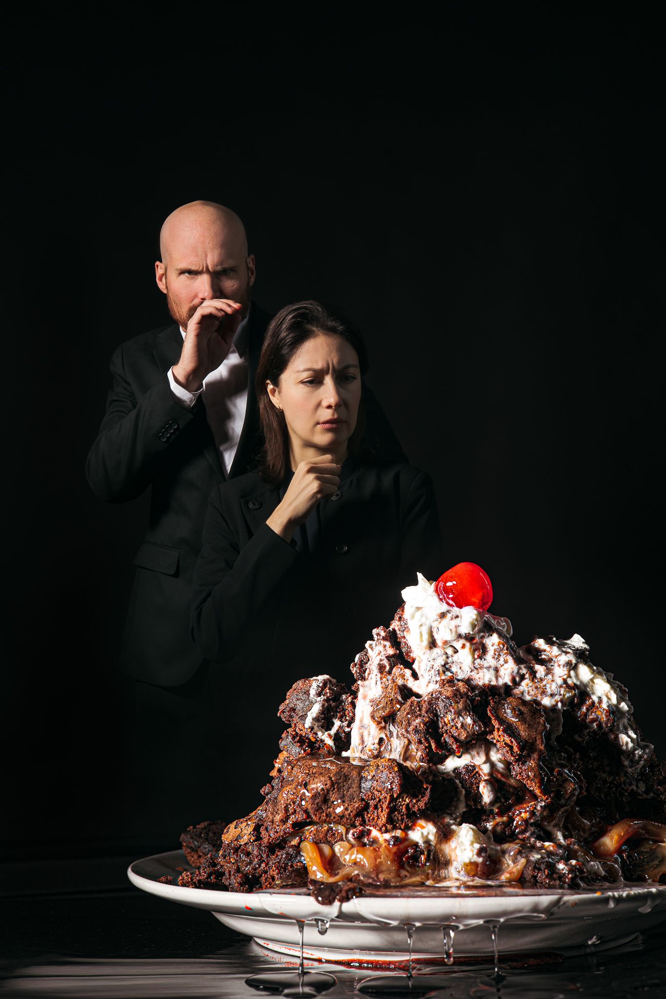 A person wearing dark clothing sits at a table with a large chocolate dessert as they eat it in dramatic lighting.
