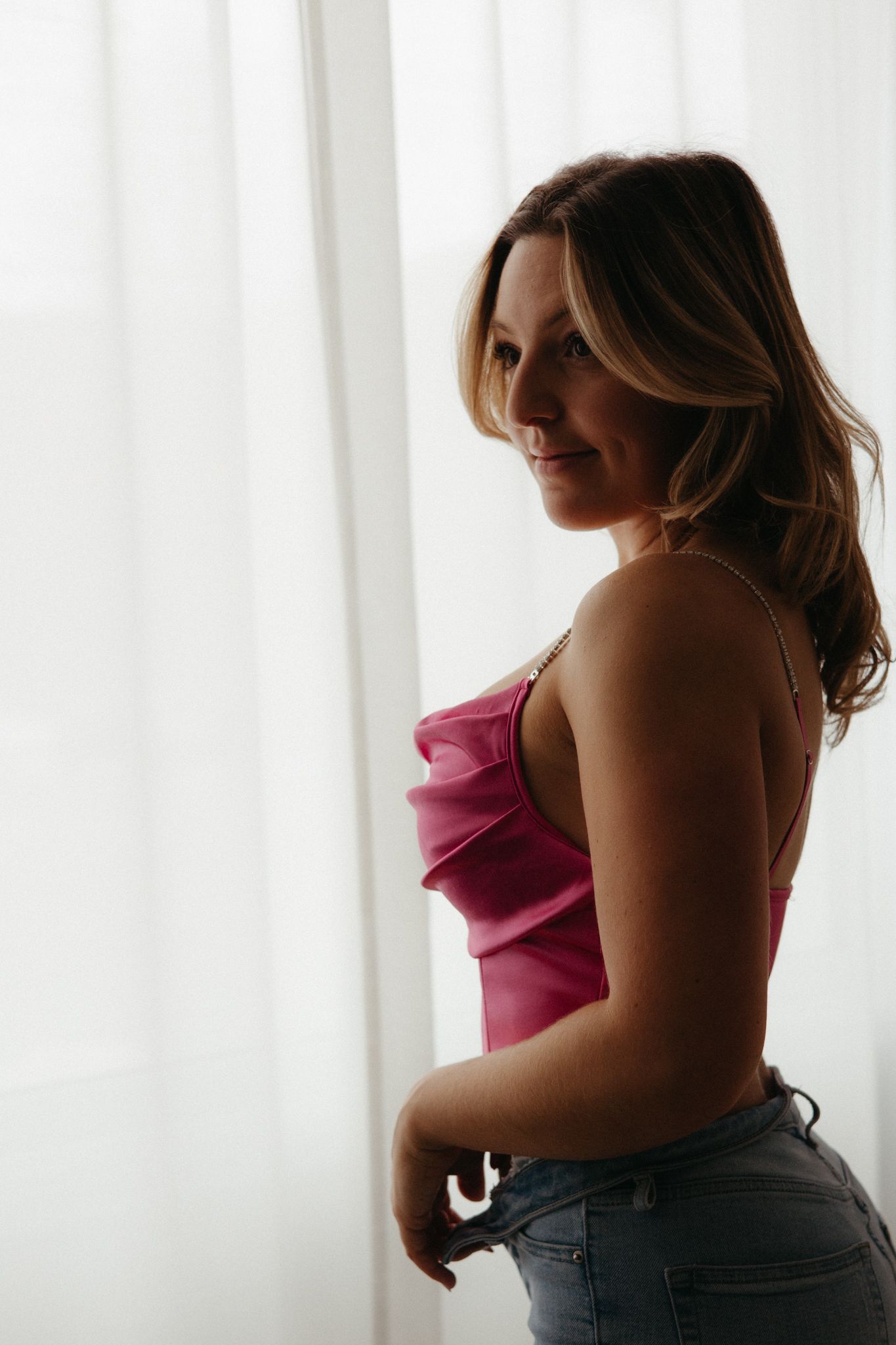 A person in a pink sleeveless top poses confidently against a white curtained background.