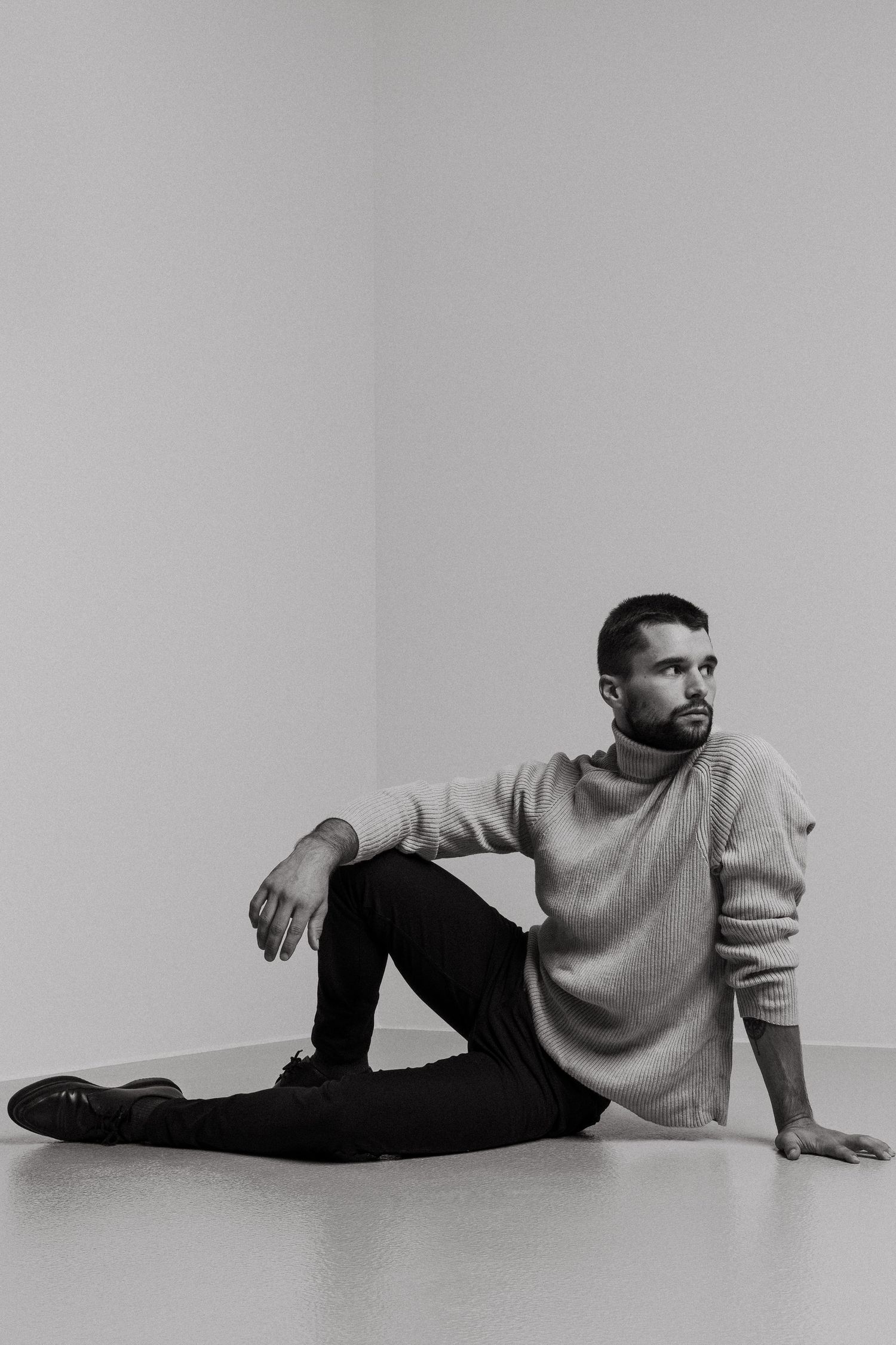 Black and white studio portrait featuring a person in a light sweater and dark pants sitting casually on the floor.