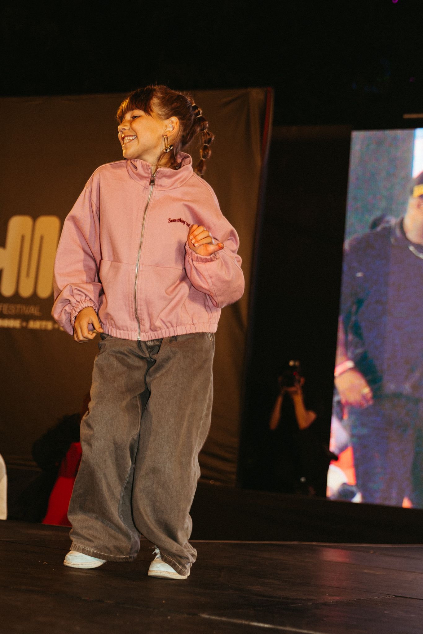 A small child in a pink top and gray pants dances across a dark stage under theatrical lighting.