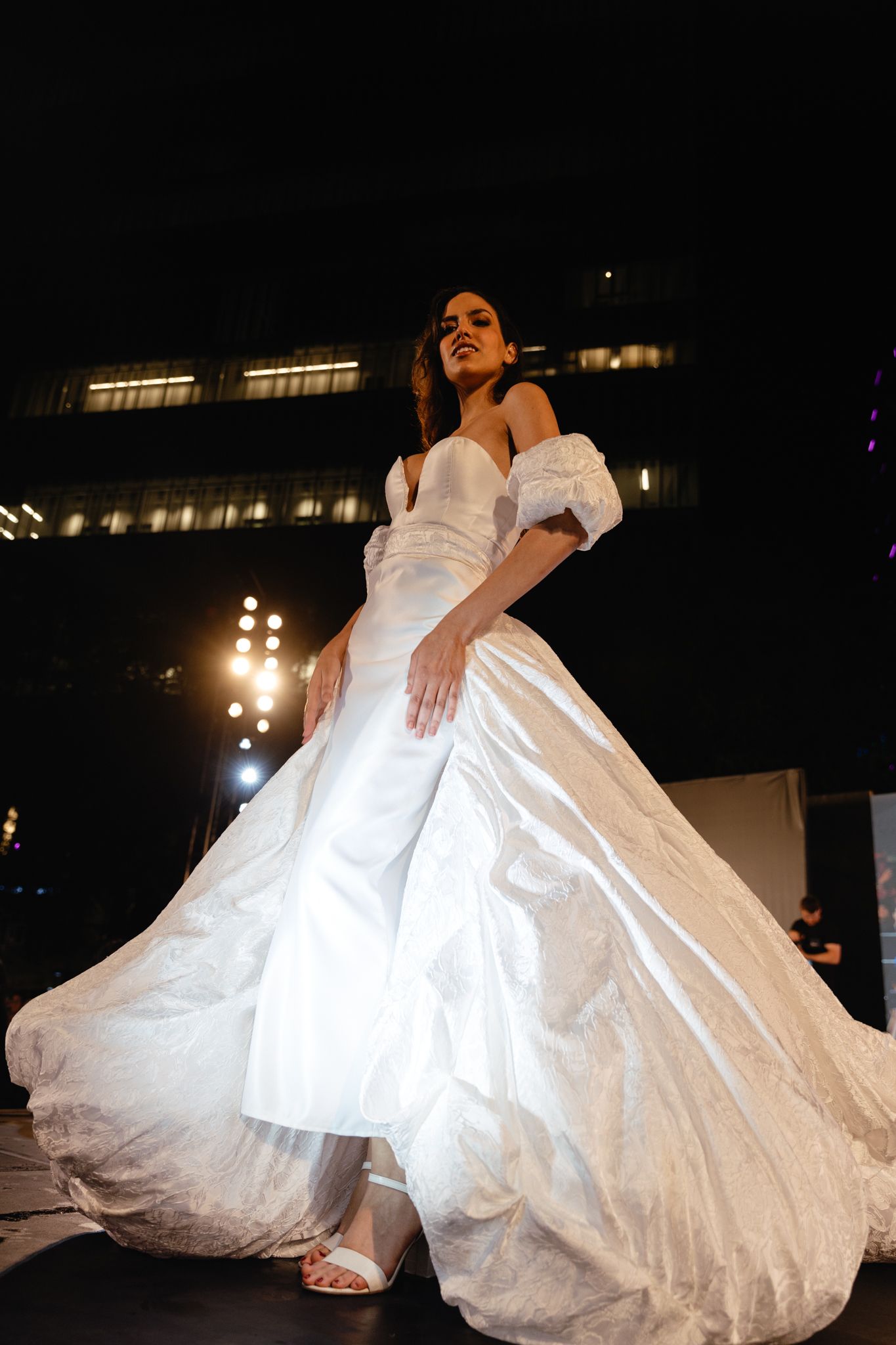 Model walks runway in dramatic off-shoulder white wedding gown with flowing train during evening fashion show.