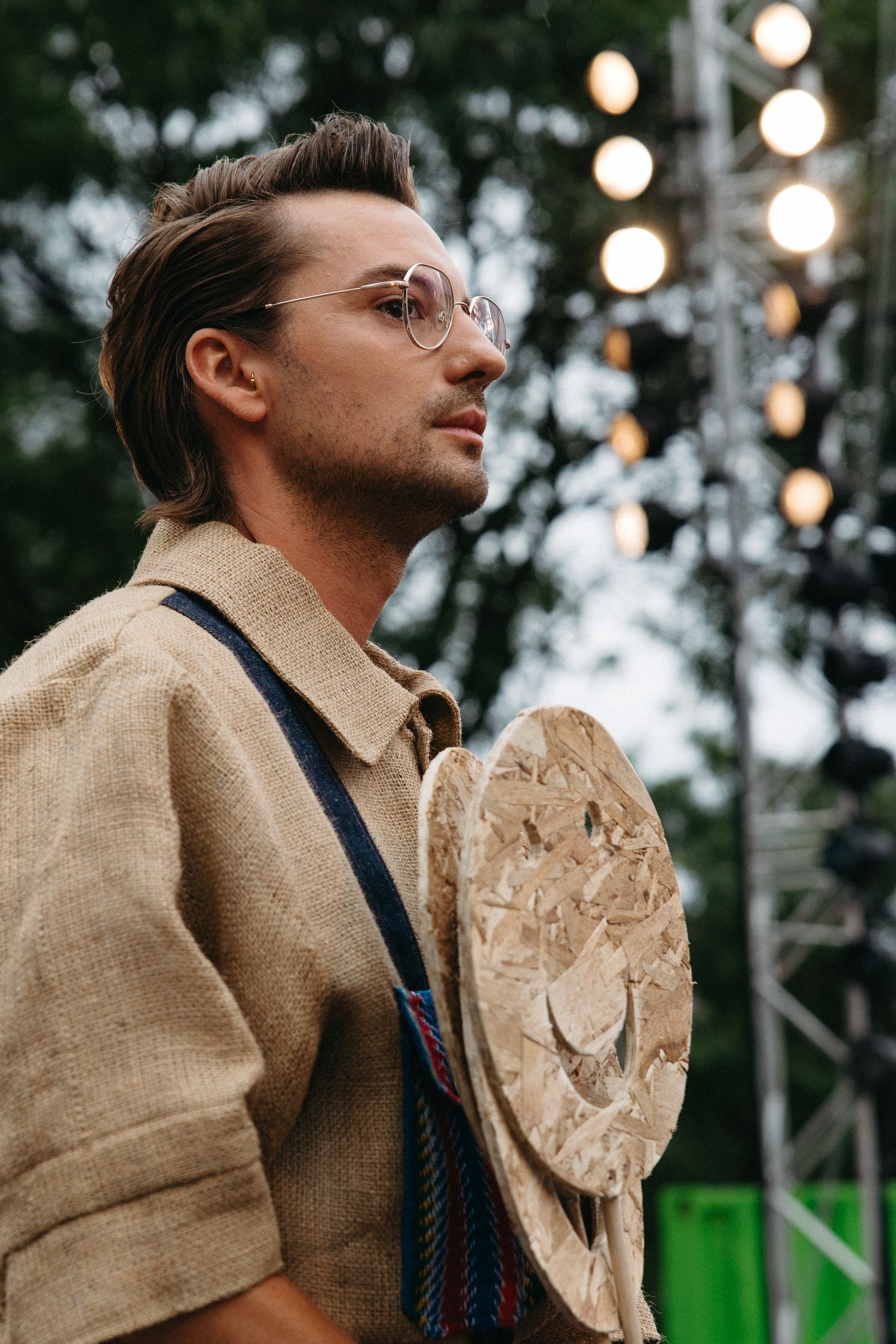 A person wearing glasses, a linen suit jacket and suspenders stands outdoors with natural lighting and bokeh in the background.
