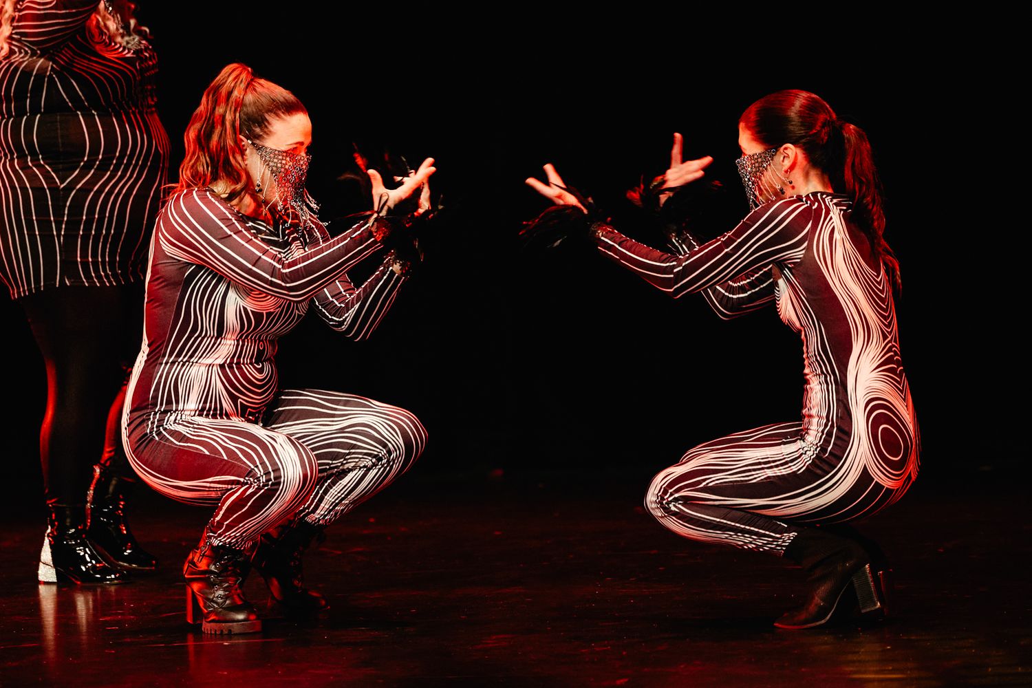 Two dancers in striped bodysuits perform a mirrored crouching pose on stage under dramatic red lighting.