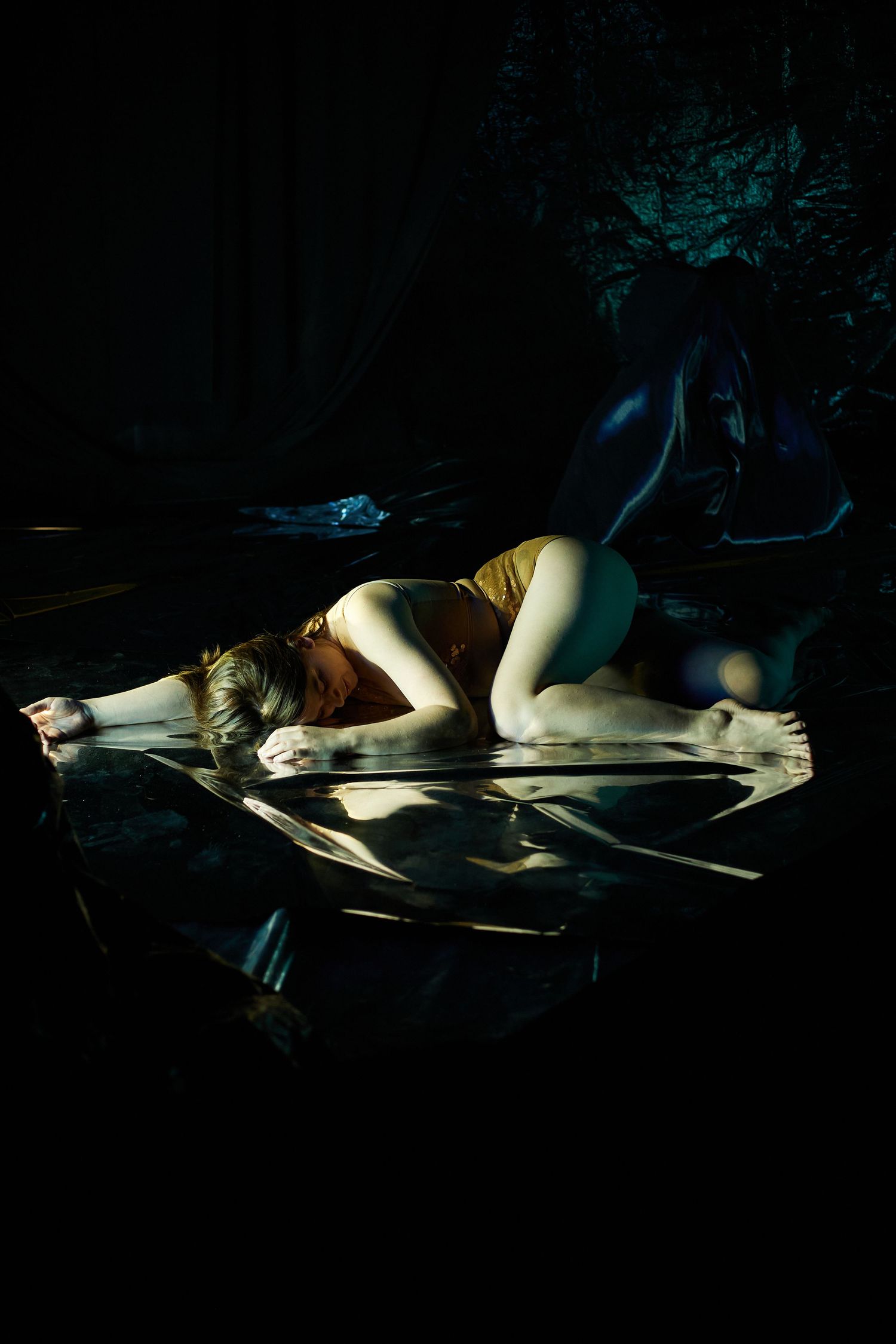 A person in yellow lies curled up on a reflective surface in a dark, moody artistic photograph.