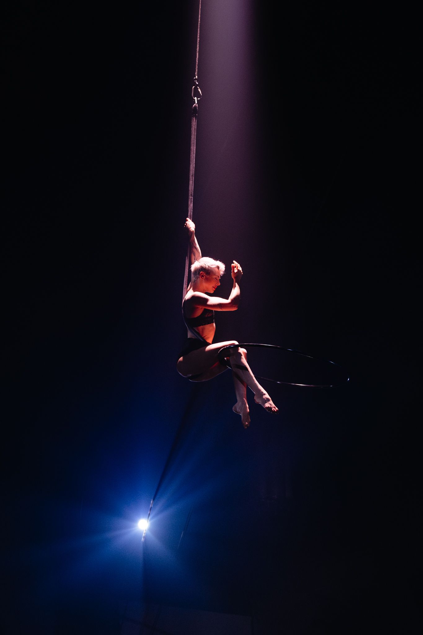 Aerial performer in red costume gracefully suspended on rope against dark background with dramatic blue spotlight.