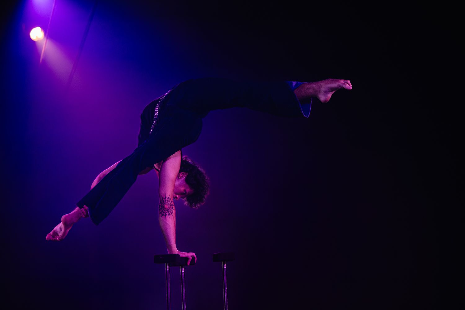 A performer executes an acrobatic pole dance routine under purple stage lighting showing impressive strength and skill.