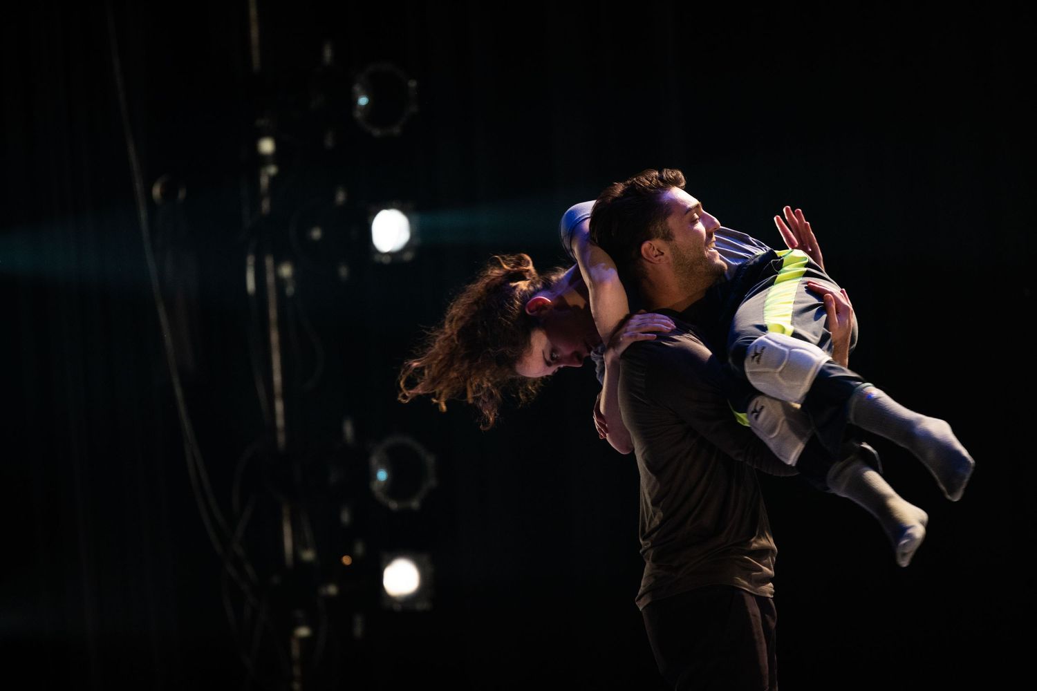 A performer on stage dances vigorously in mid-air with dramatic lighting and dark background during a show.