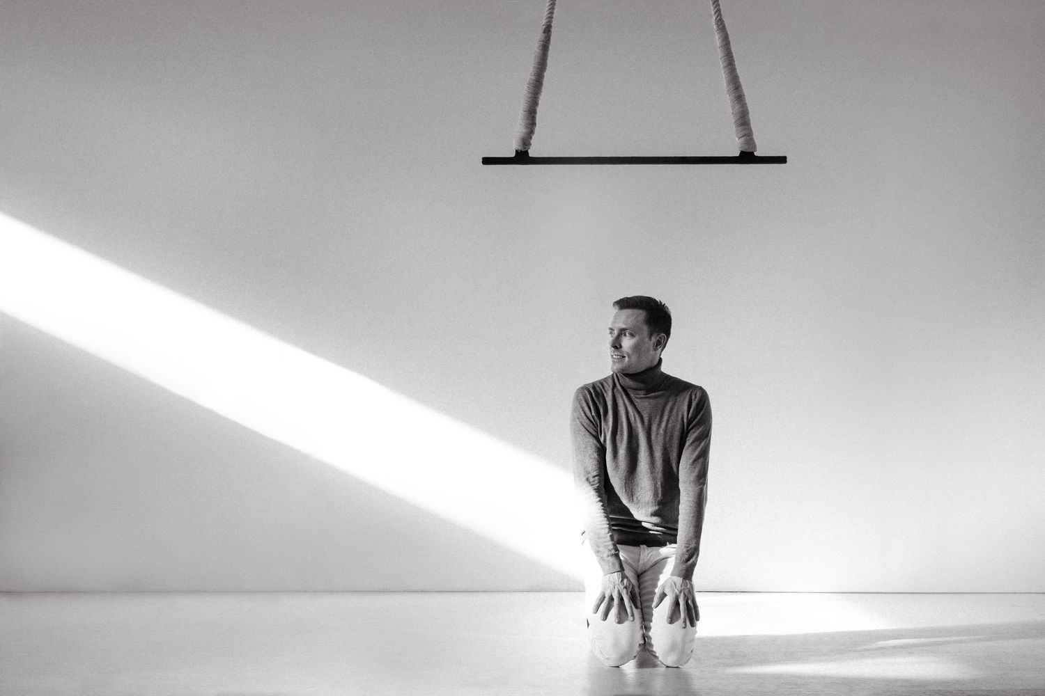 A person kneels in a minimalist white room with dramatic diagonal beam of light streaming across the scene.