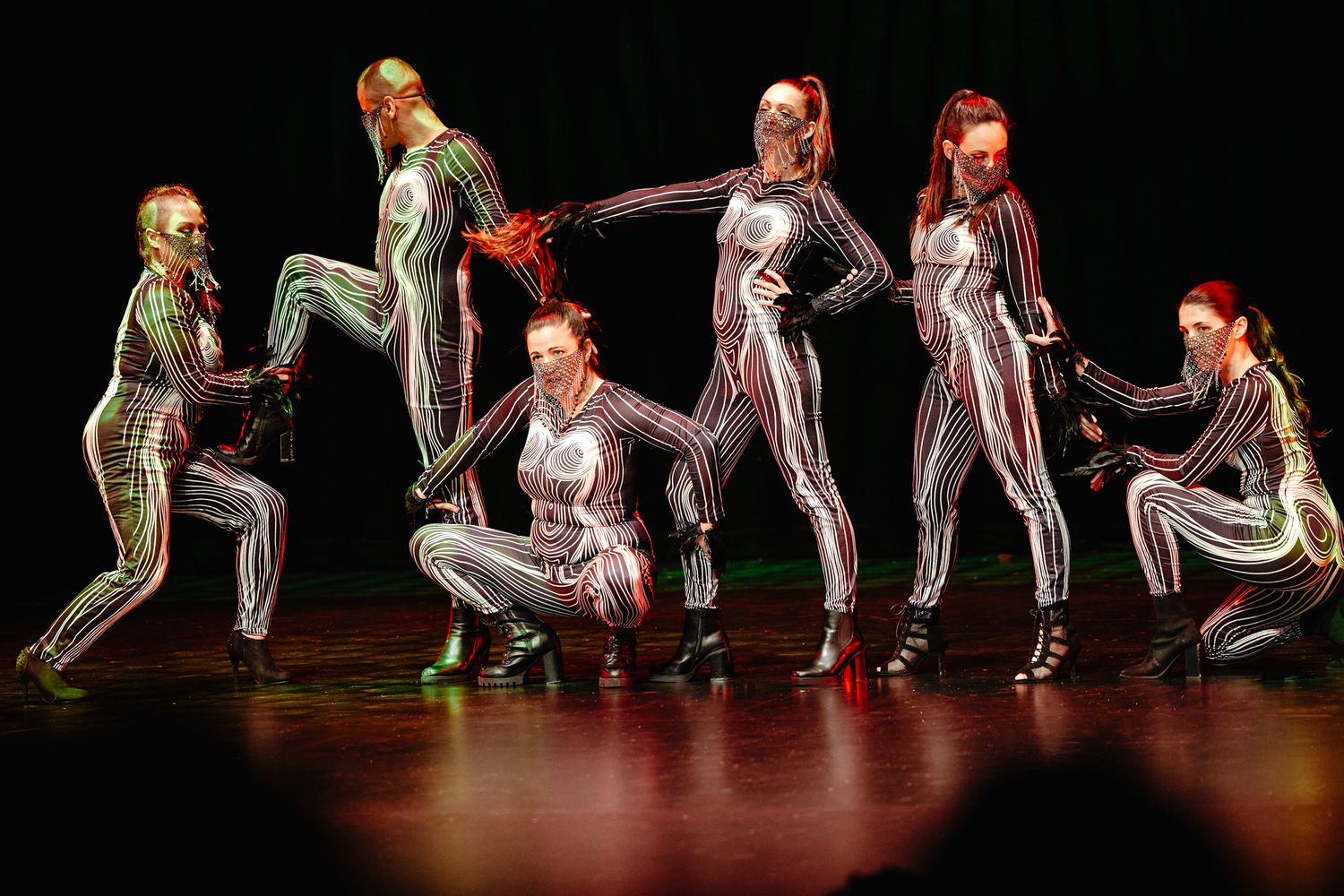 Multiple dancers in bright workout attire perform a synchronized movement sequence on a dark stage.