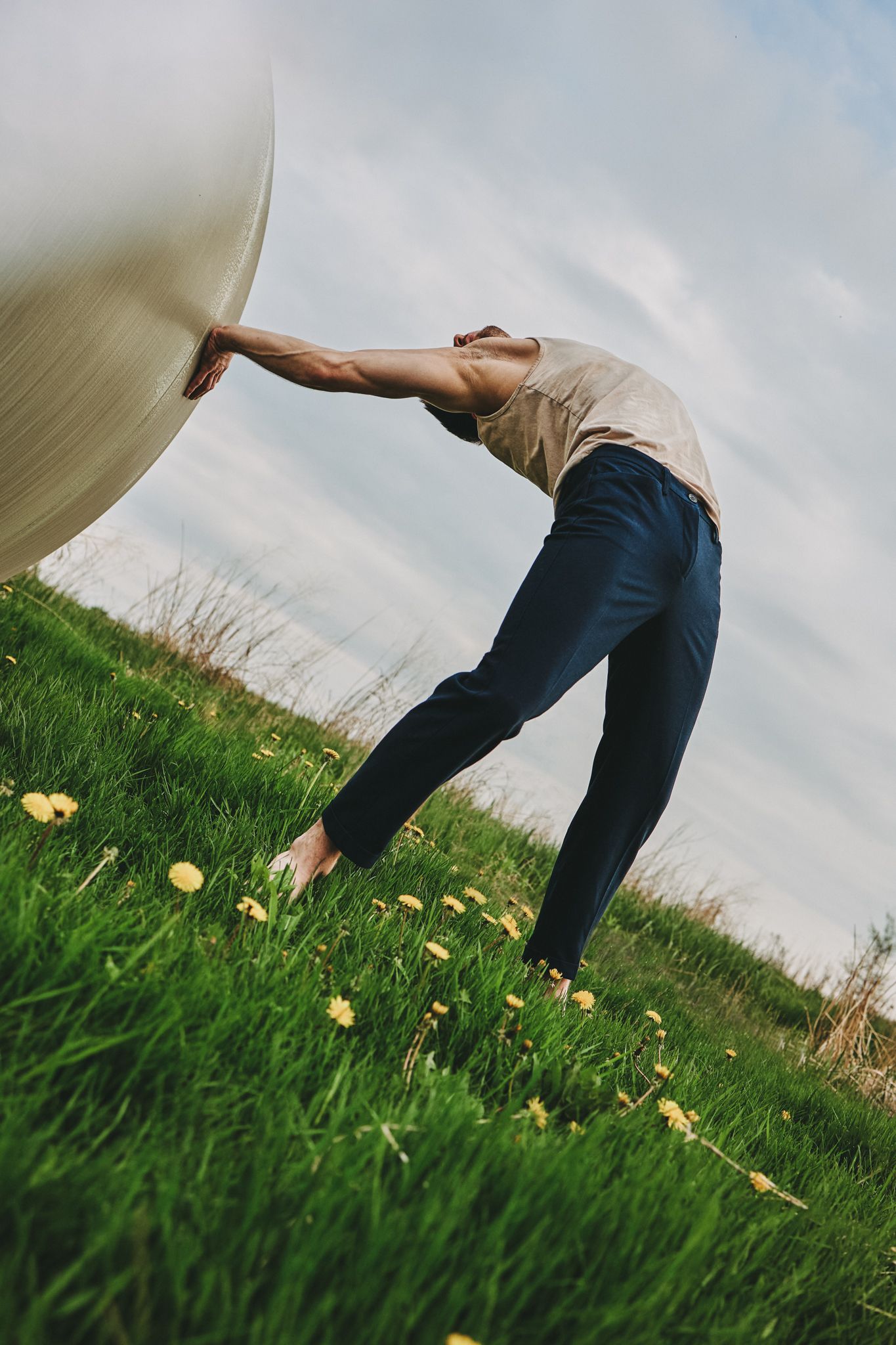 Person in beige top and jeans bends backwards in grassy field with dandelions under cloudy sky.