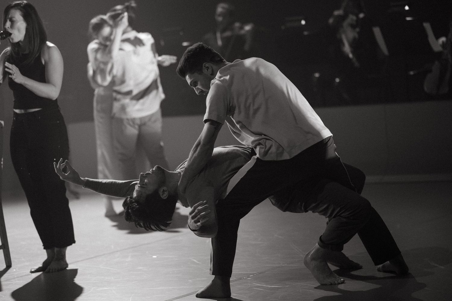 Dancers performing contemporary dance moves in dramatic black and white on a dimly lit stage.