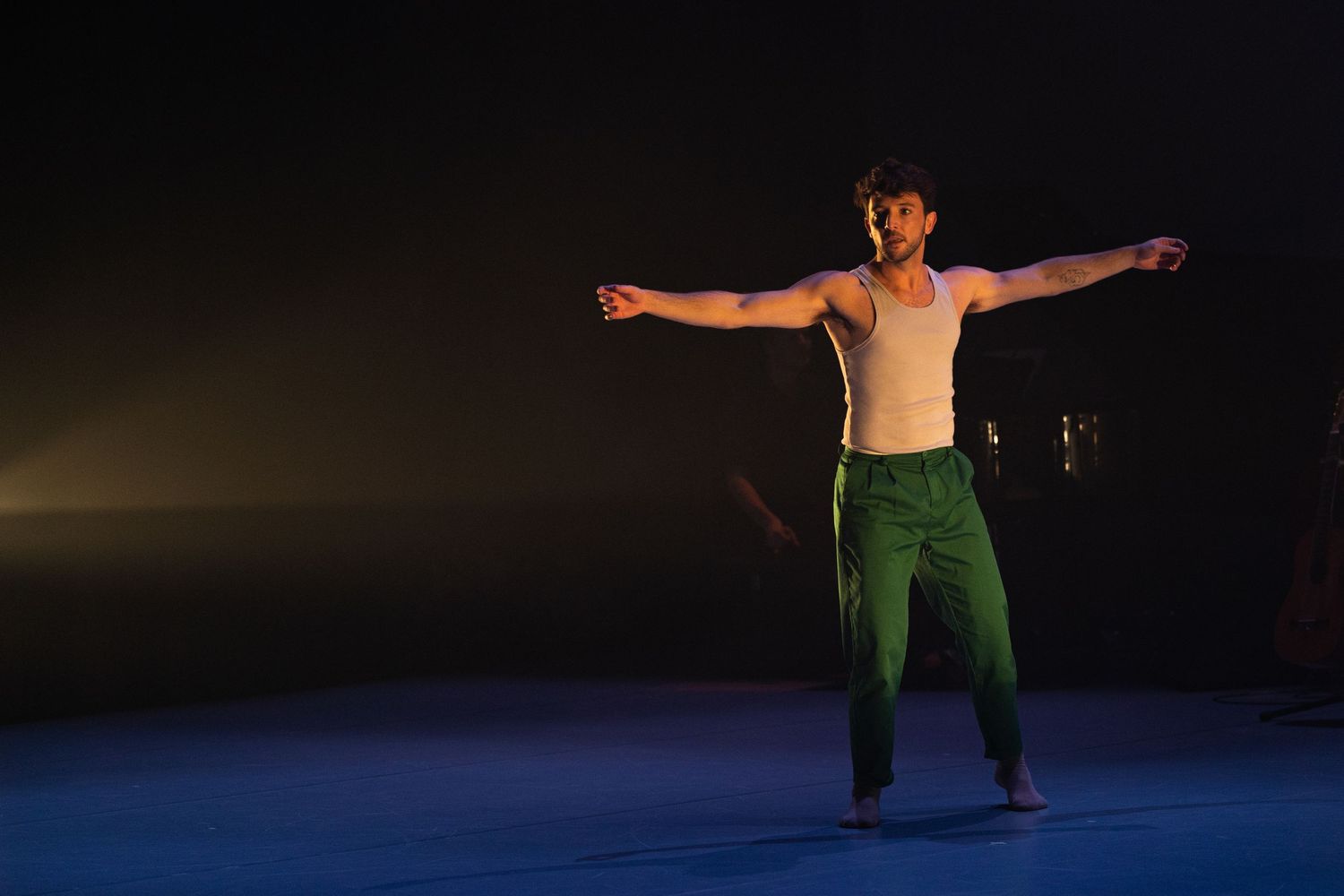A male dancer in green pants performs dynamic movements on a dark stage, creating multiple exposure effect.