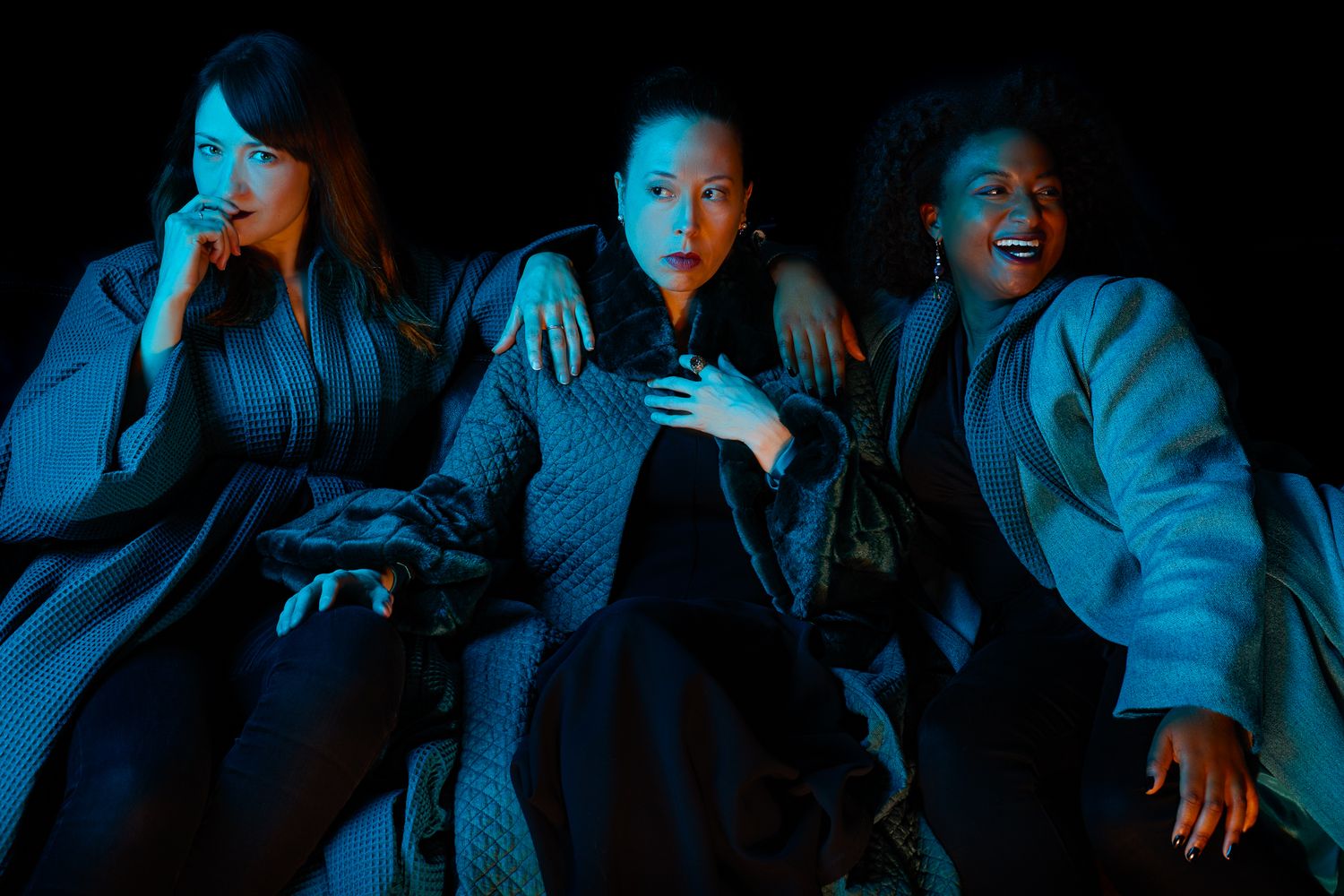 Three people react dramatically with shocked expressions while sitting together under moody blue lighting.