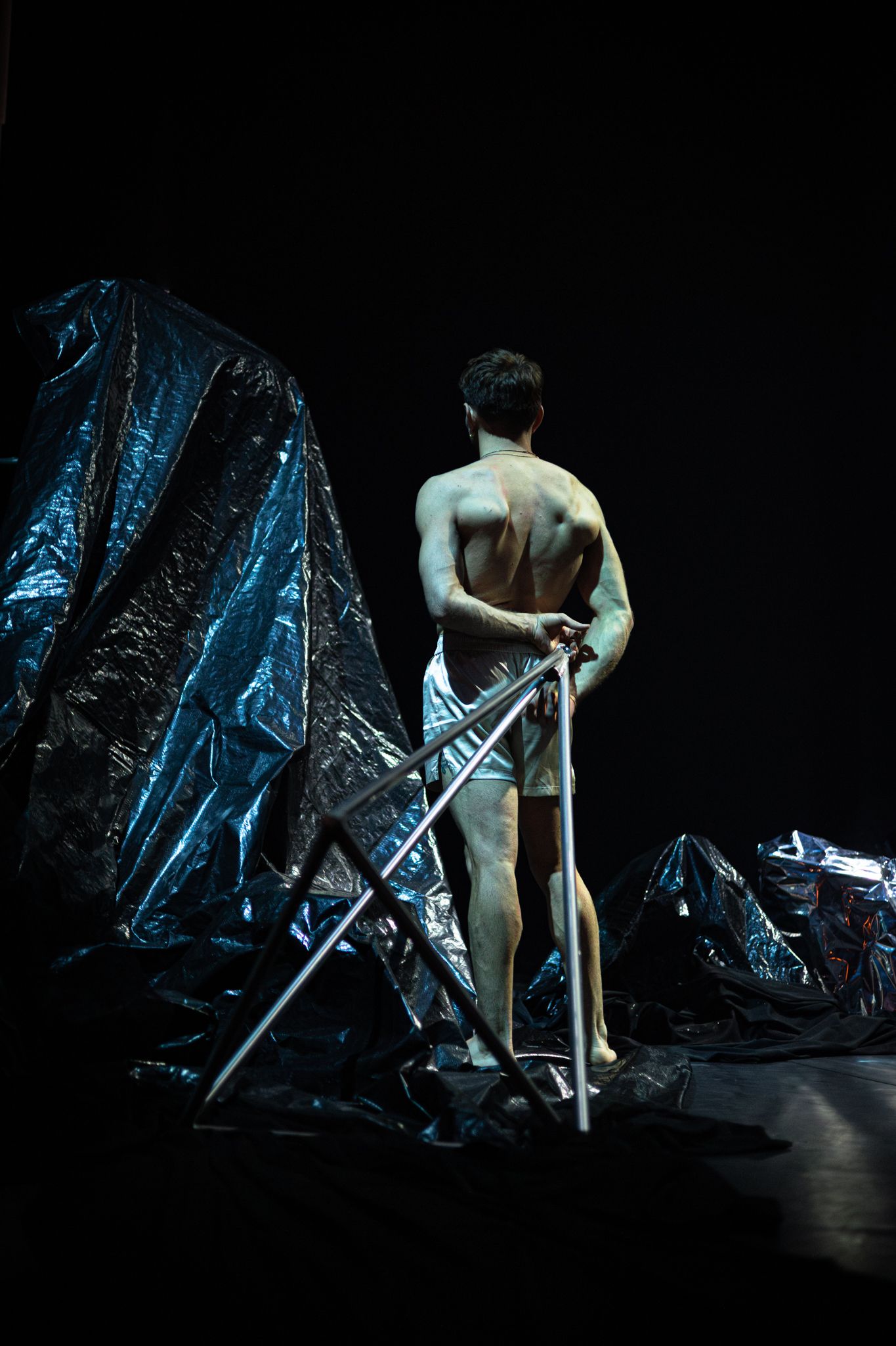 A shirtless performer stands on stage with crutches next to metallic blue curtains in dramatic theatrical lighting.