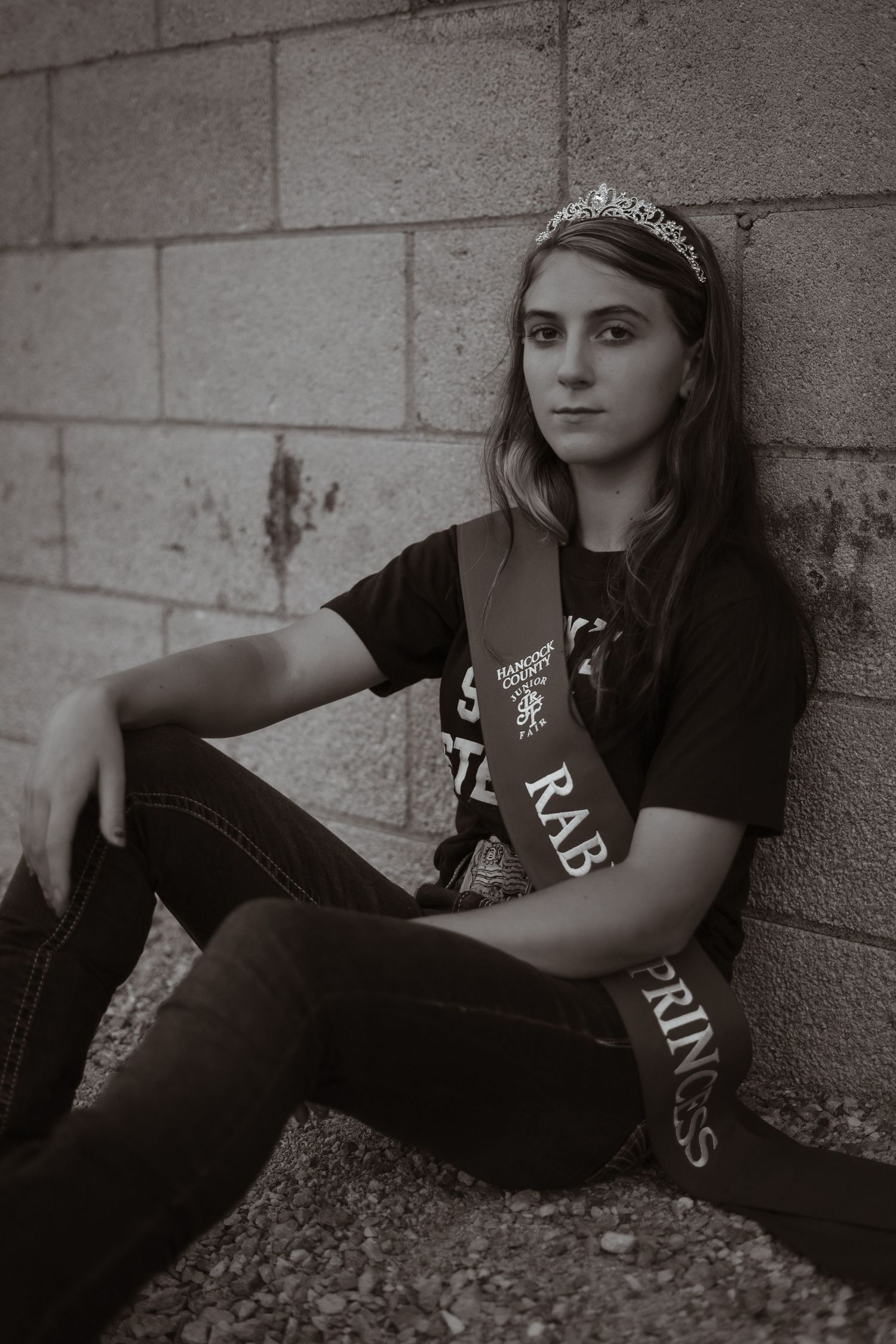 A young person in casual attire sits against a concrete wall wearing a tiara and band t-shirt.