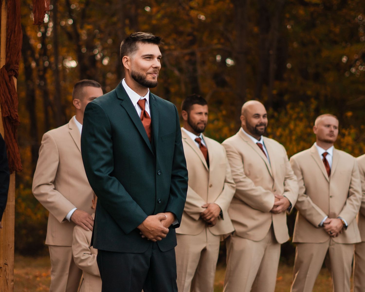 Groom in dark green suit smiles at altar with groomsmen in tan suits standing behind him outdoors.