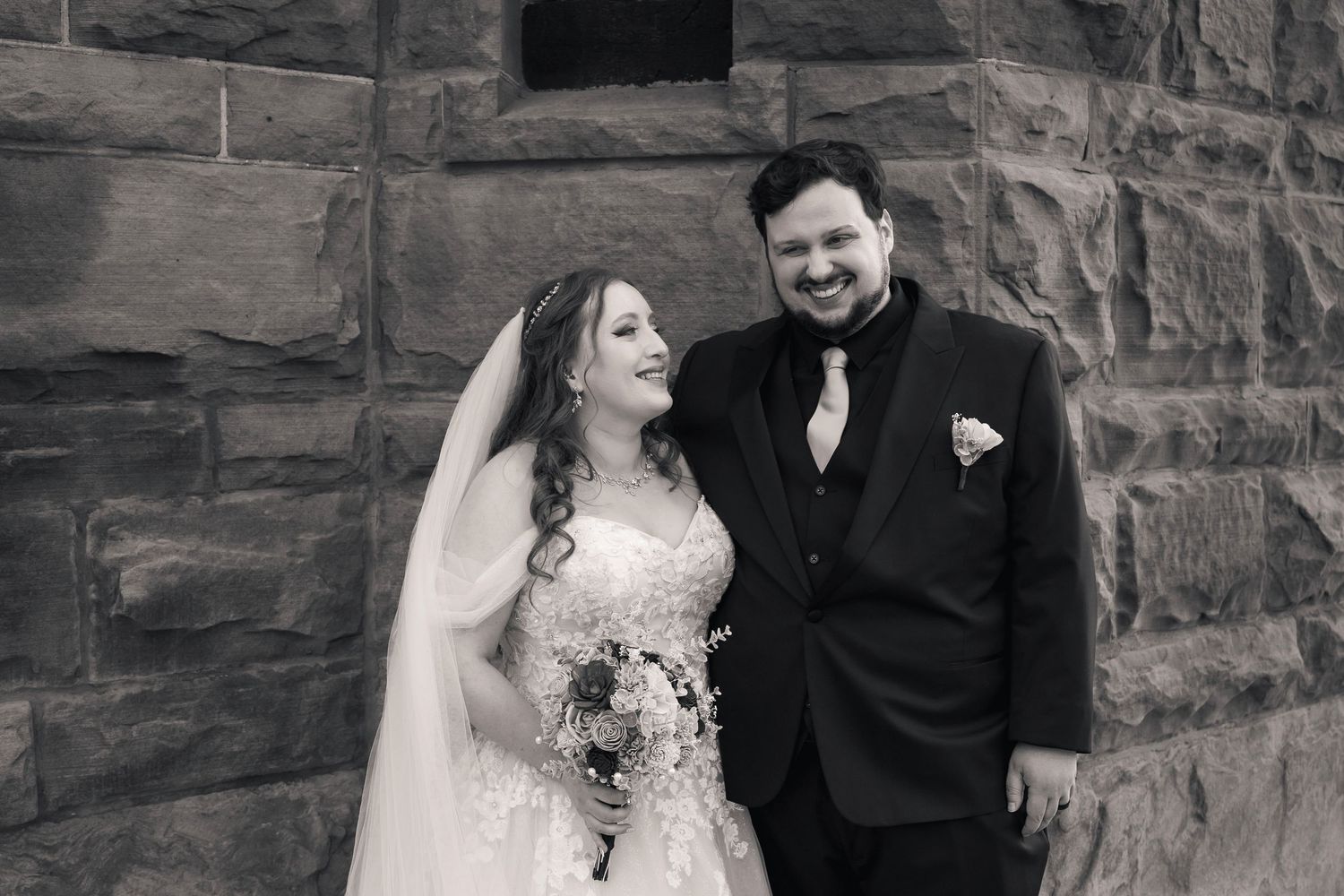Black and white wedding photo of smiling bride in veil holding bouquet and groom in suit against stone wall.