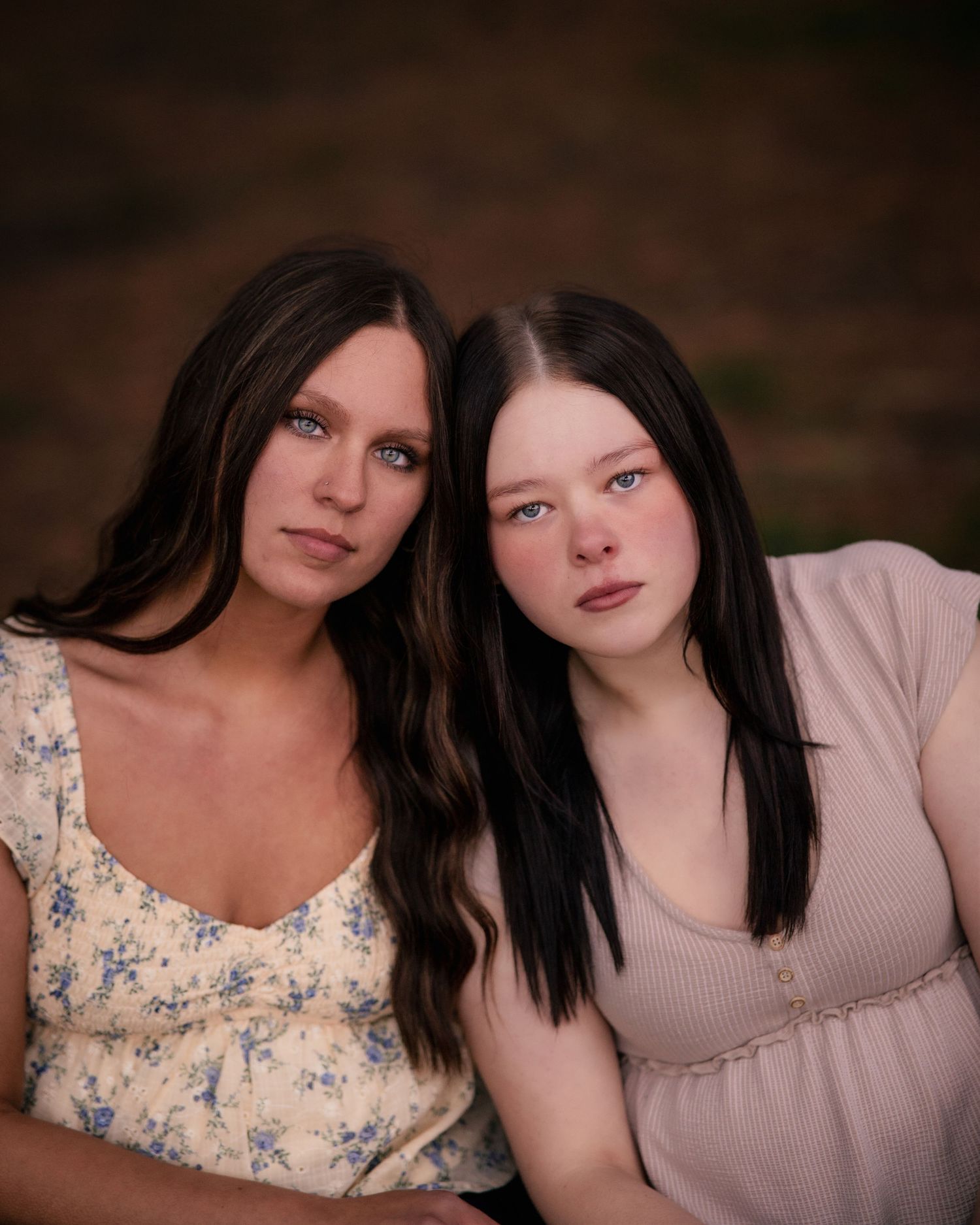 Two dark-haired women pose closely together outdoors, wearing floral and pink tops in a warm portrait.