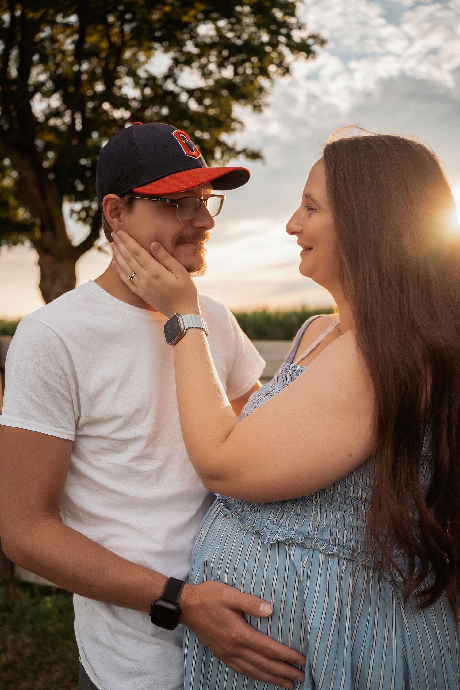 A couple shares a tender moment outdoors at sunset, with the woman touching the man's face as he holds her pregnant belly.