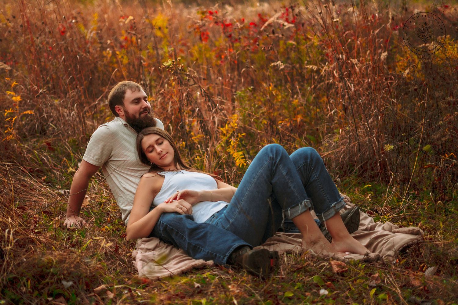 Couple relaxes on blanket in tall autumn grass during sunset photoshoot.