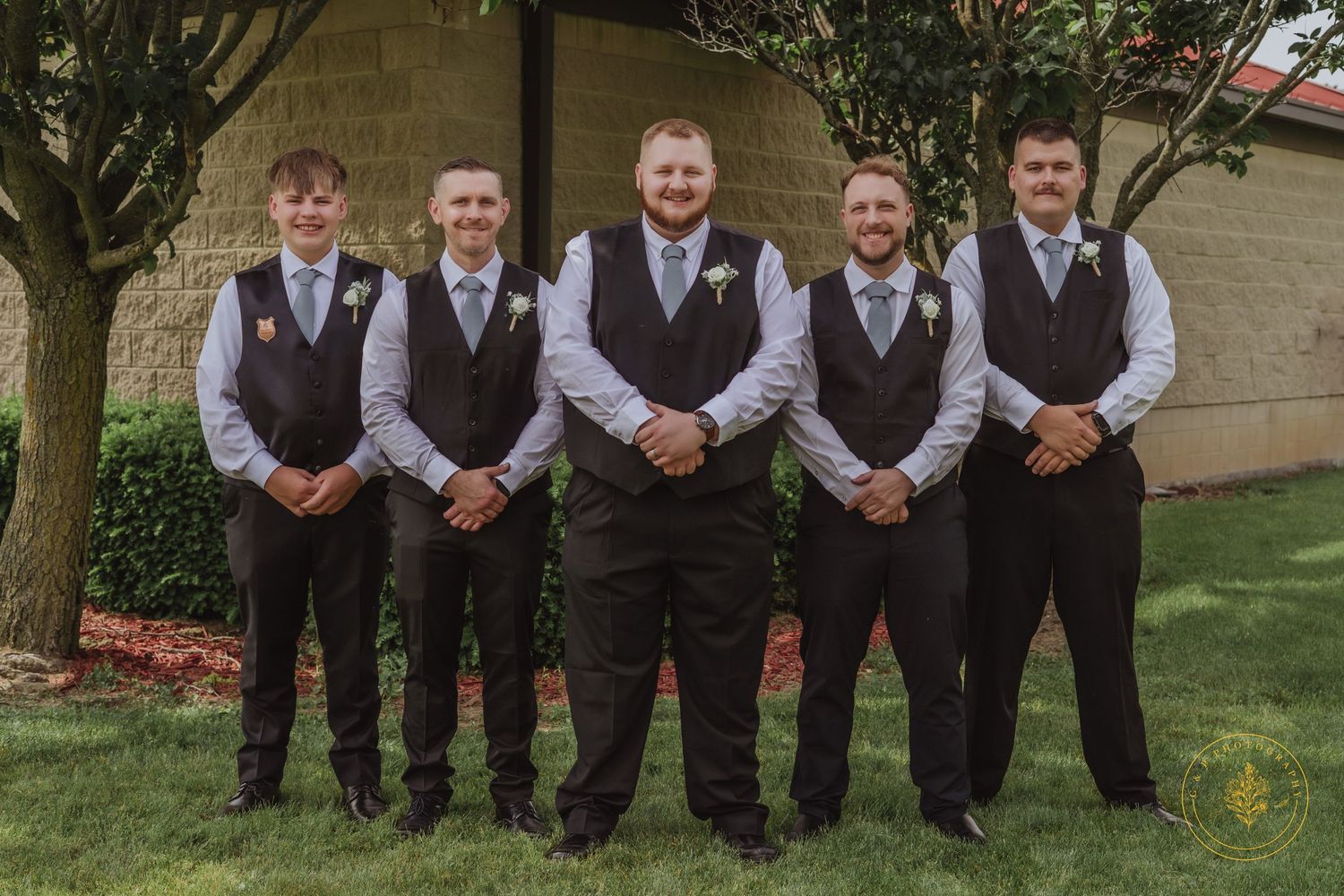 A wedding party wearing matching brown vests and ties poses together outdoors.