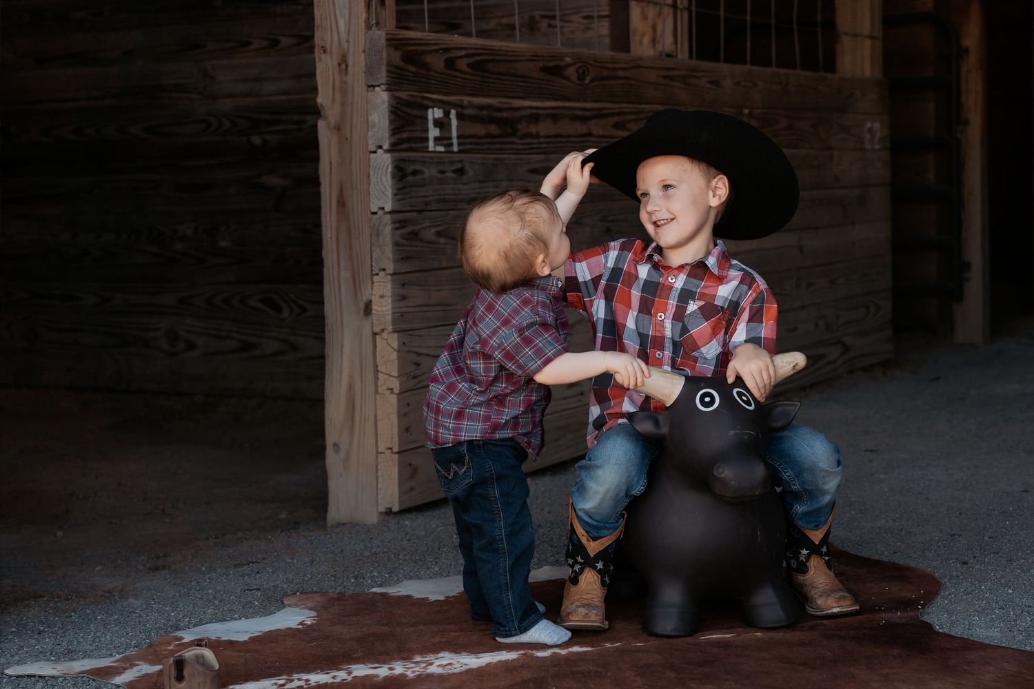 Two young boys in cowboy hats and plaid shirts play on a toy horse in a rustic barn setting.