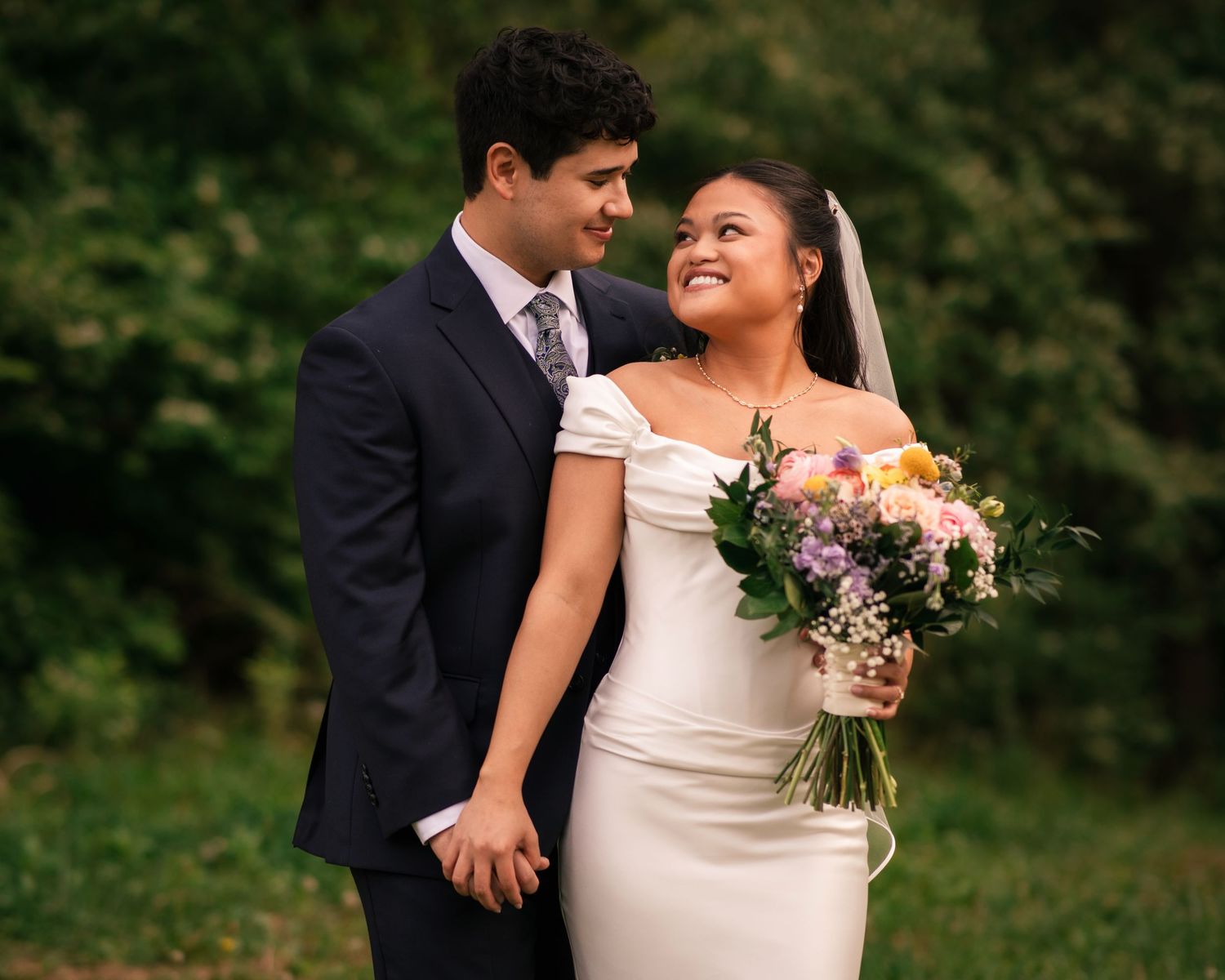Bride and groom holding hands and smiling at each other outdoors, bride holding colorful bouquet in white wedding dress.
