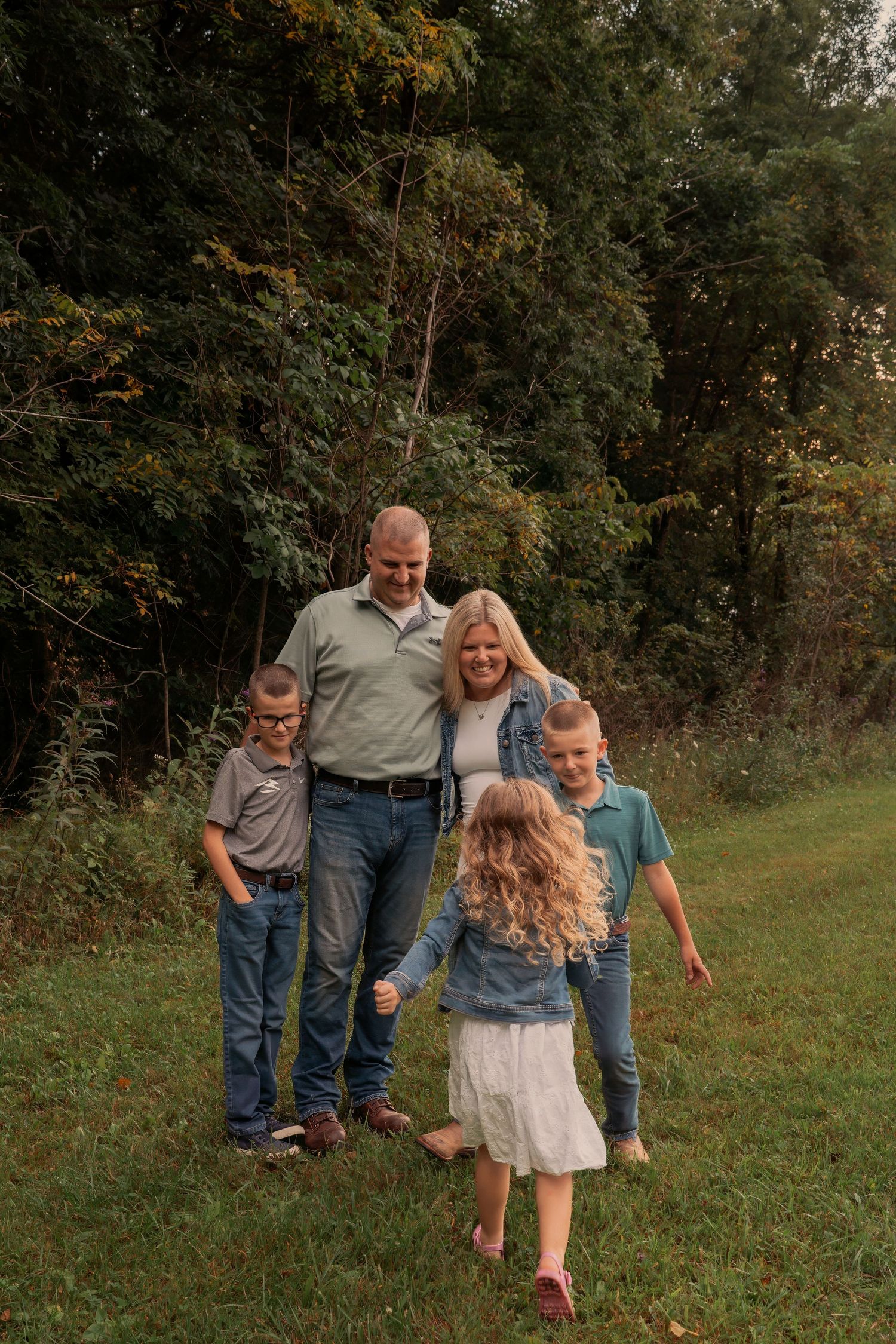 A happy family of five smiles outdoors in a lush green field, with two parents and three children enjoying a casual moment.