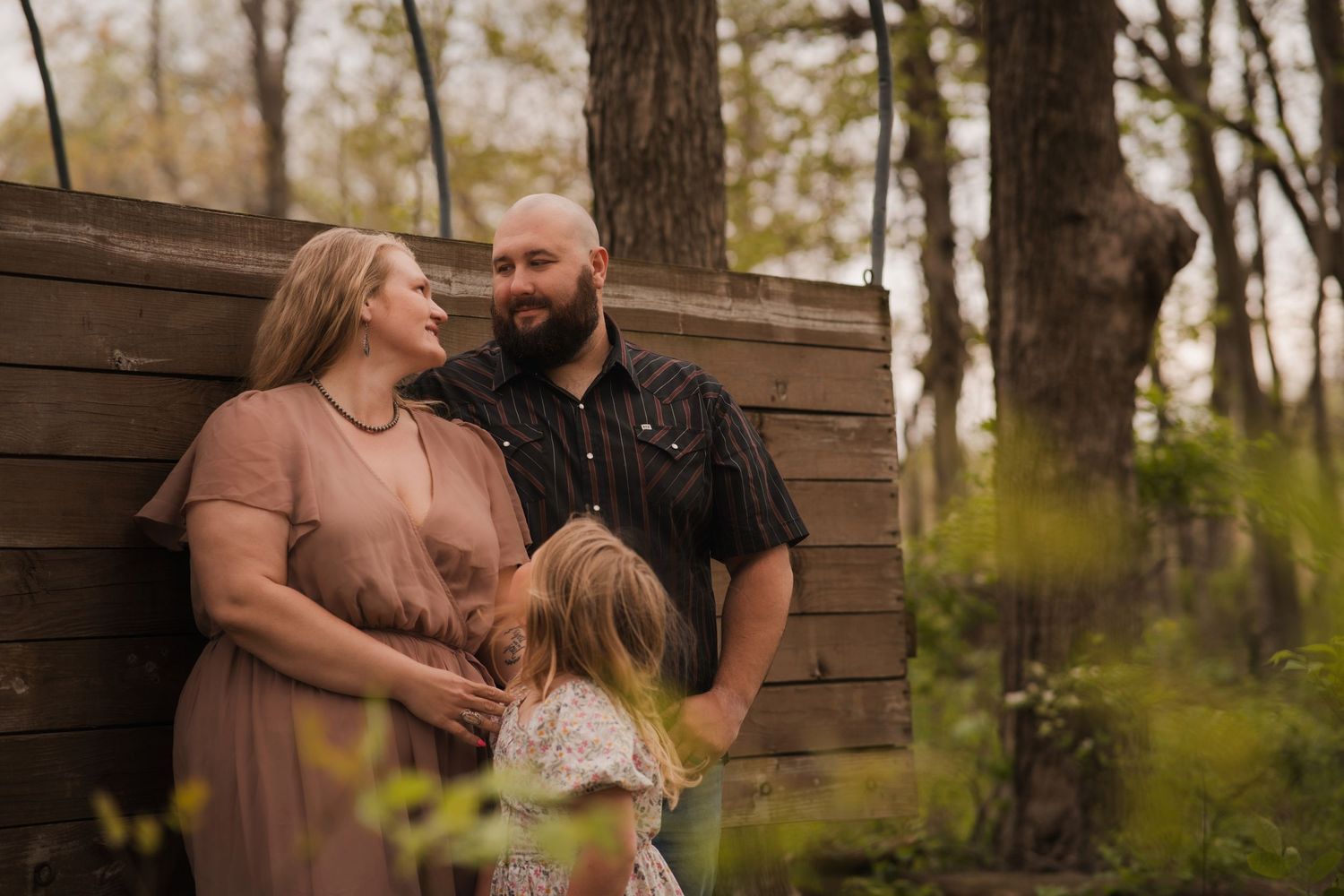 Pregnant couple and young child share a tender moment outdoors near a wooden fence surrounded by trees.