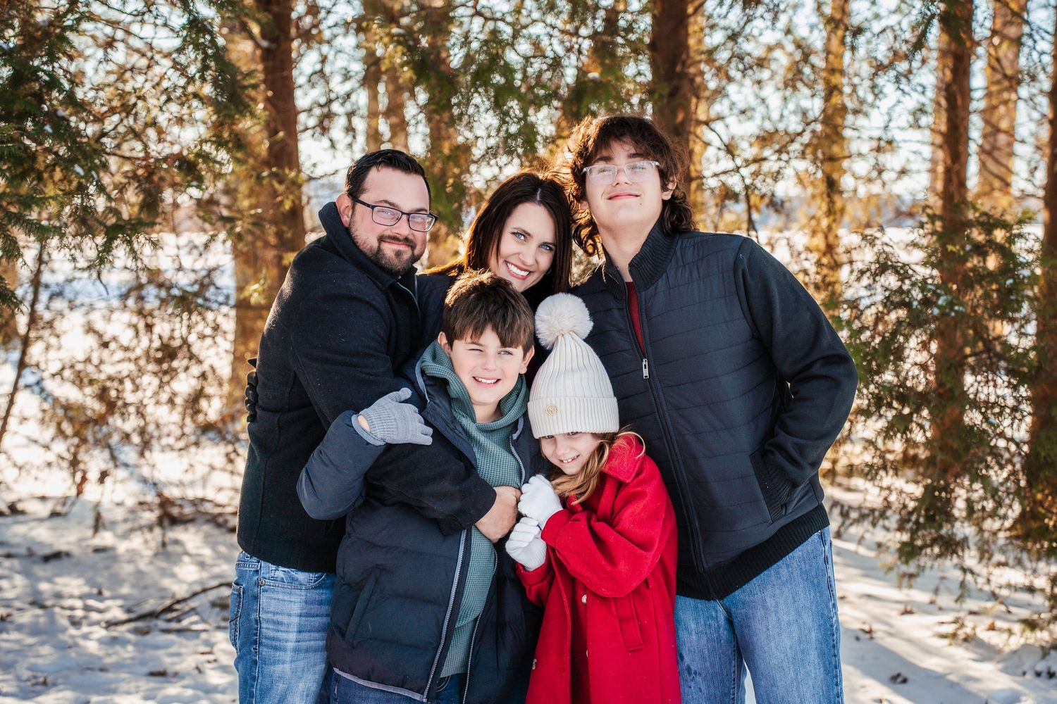 A happy family of five poses together for a winter portrait in the snow among trees.