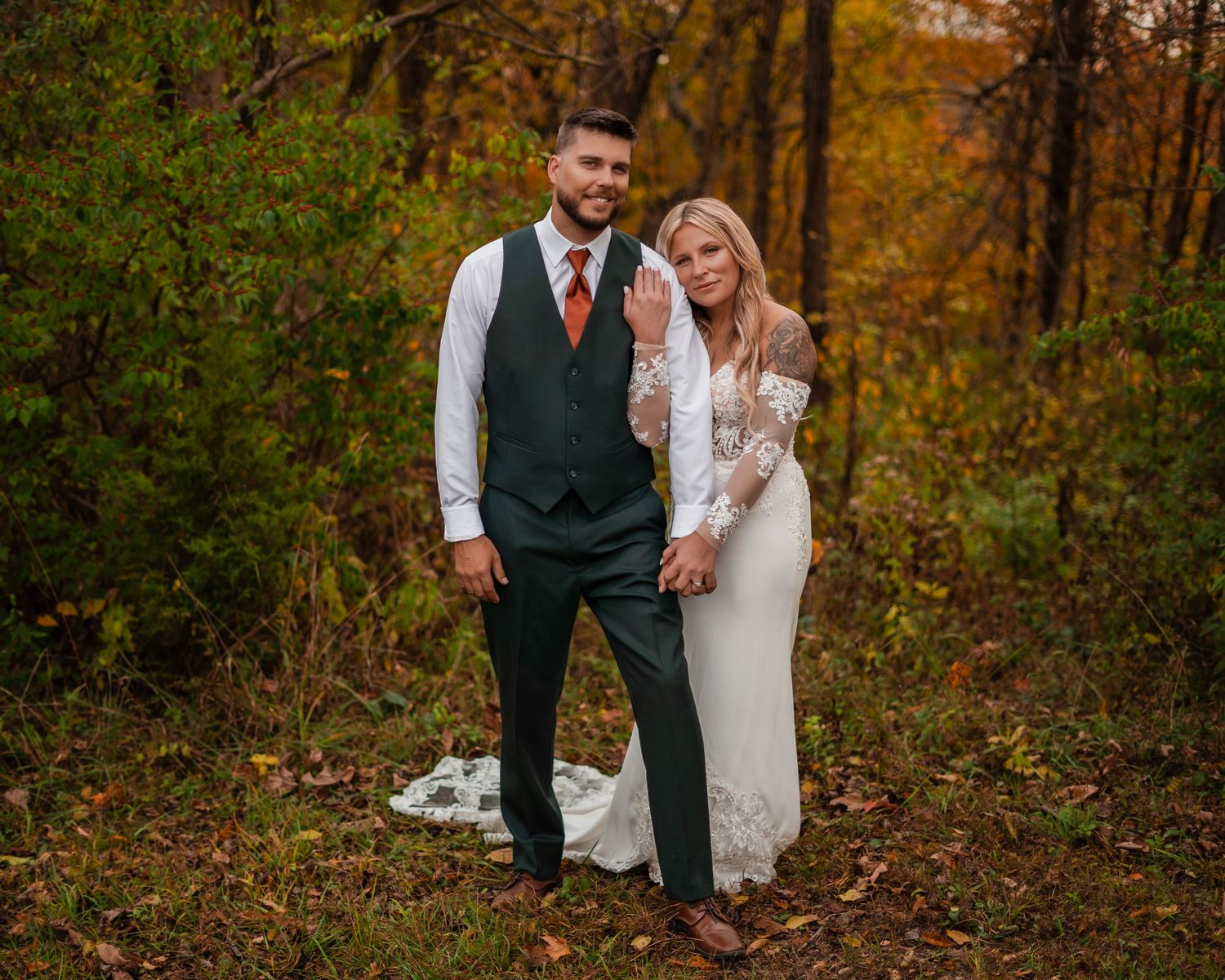 A newlywed couple stands together on fallen leaves in an autumn forest setting.