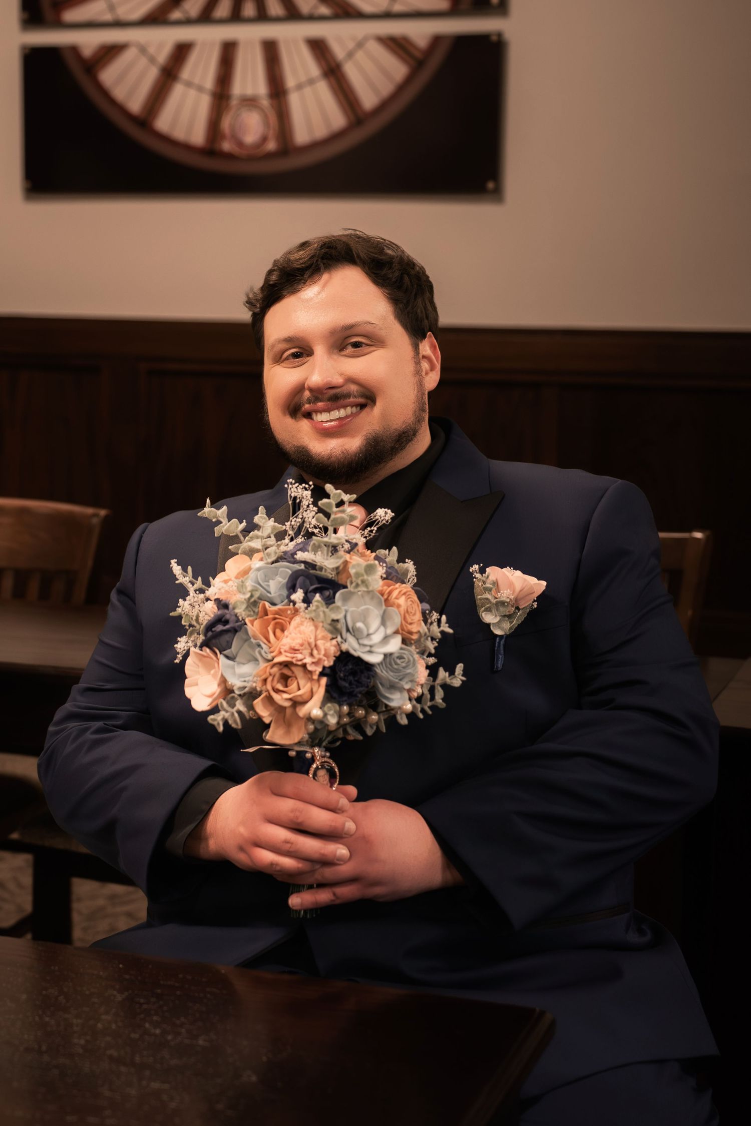 Smiling groom in navy suit holding blue and peach wedding bouquet with boutonniere, seated at wooden table.