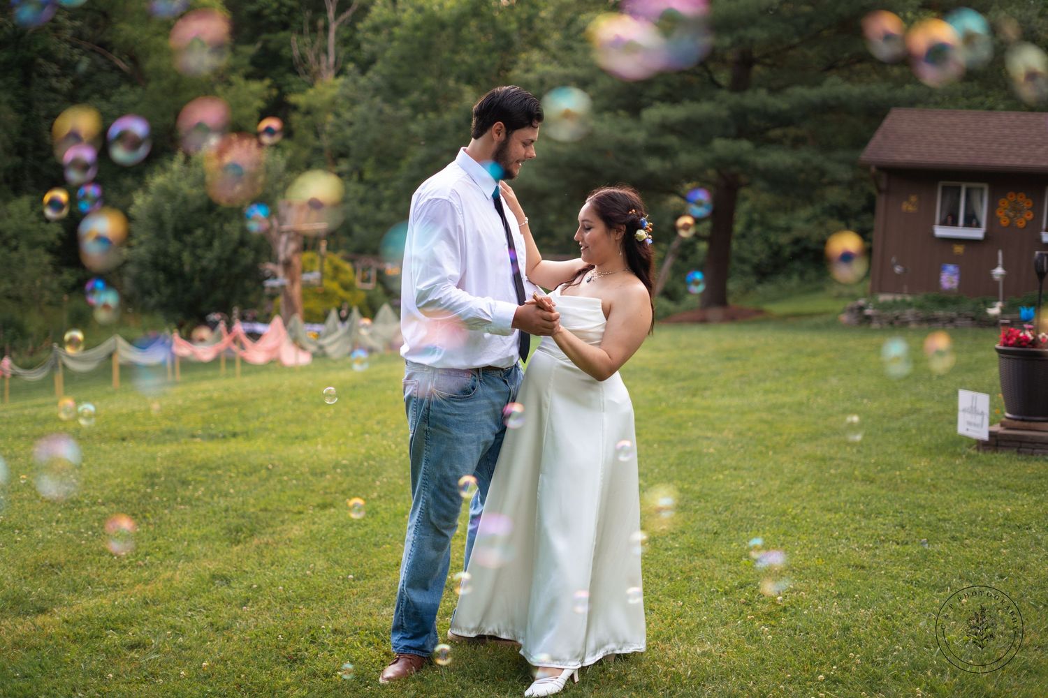 Couple shares intimate moment during outdoor wedding ceremony with floating bubbles in background.