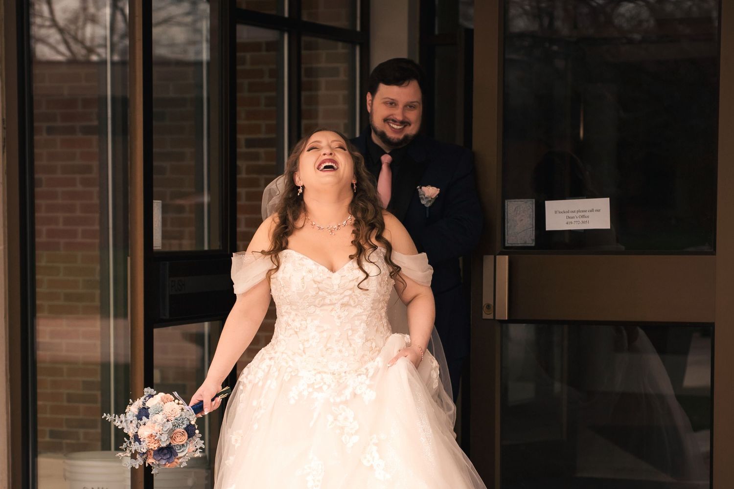 Joyful bride and groom exiting building, bride laughing in lace ball gown holding bouquet on wedding day.