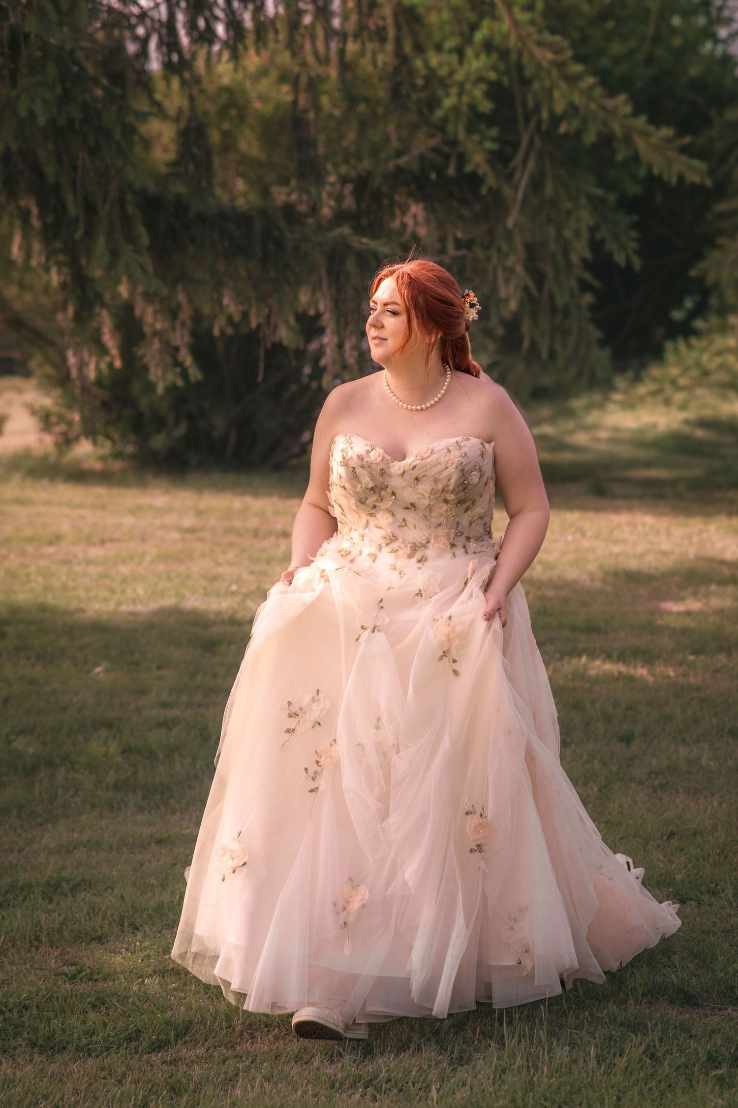 A red-haired bride in a blush floral ballgown with pearl necklace poses outdoors in golden hour light.