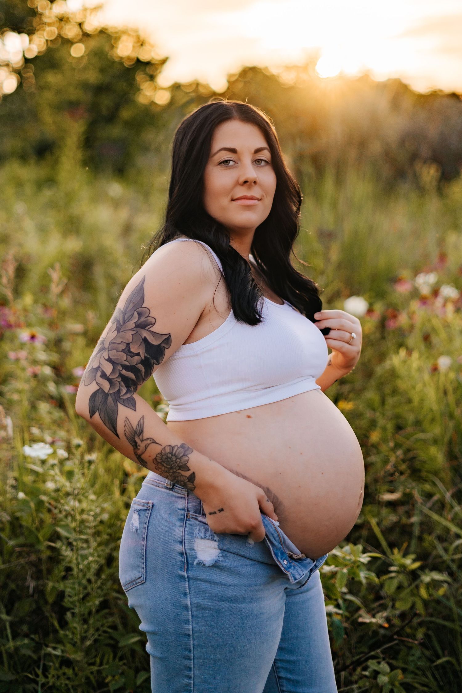Maternity photo in wildflower field at sunset showing woman in jeans and white top.