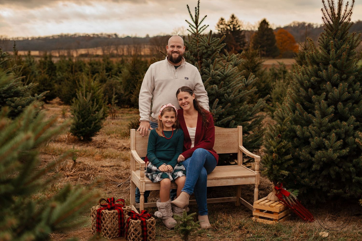 A family sitting on a wooden bench at a Christmas tree farm with wrapped presents.