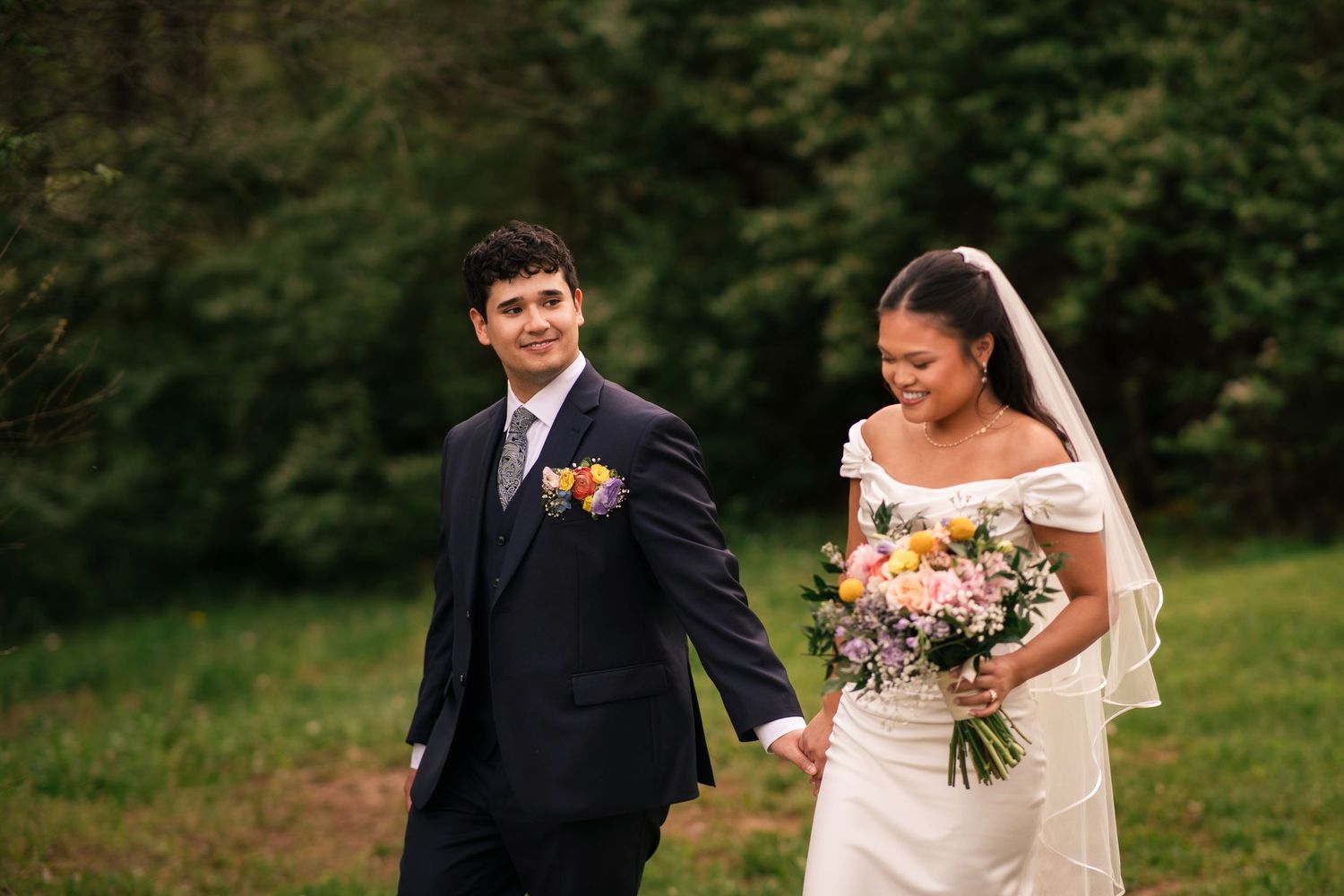 A smiling bride in a white off-shoulder gown holds a pastel bouquet while walking with a groom in a navy suit outdoors.