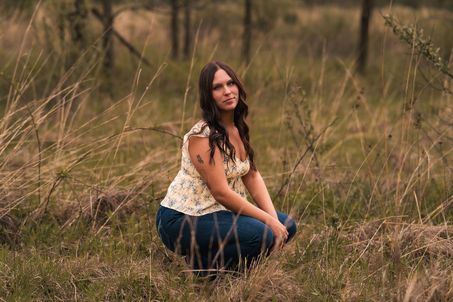 Young woman with dark hair sitting in tall grass field wearing floral top and jeans outdoors.