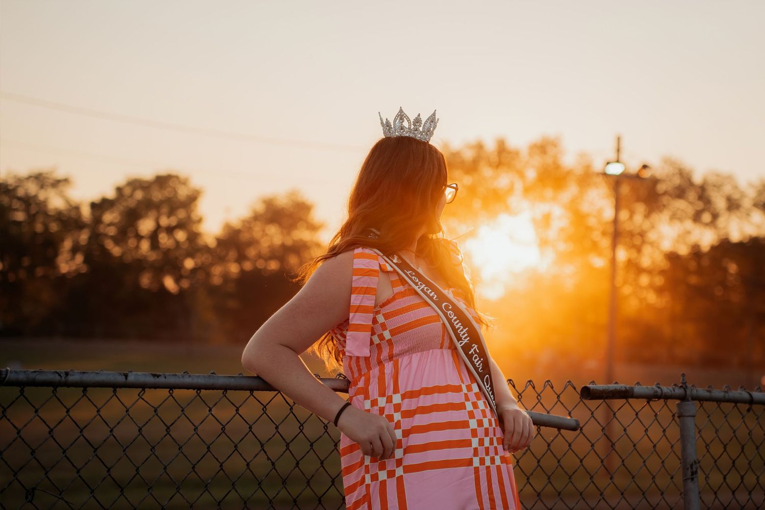 Young woman wearing a crown and orange plaid dress stands by a chain-link fence at golden sunset.