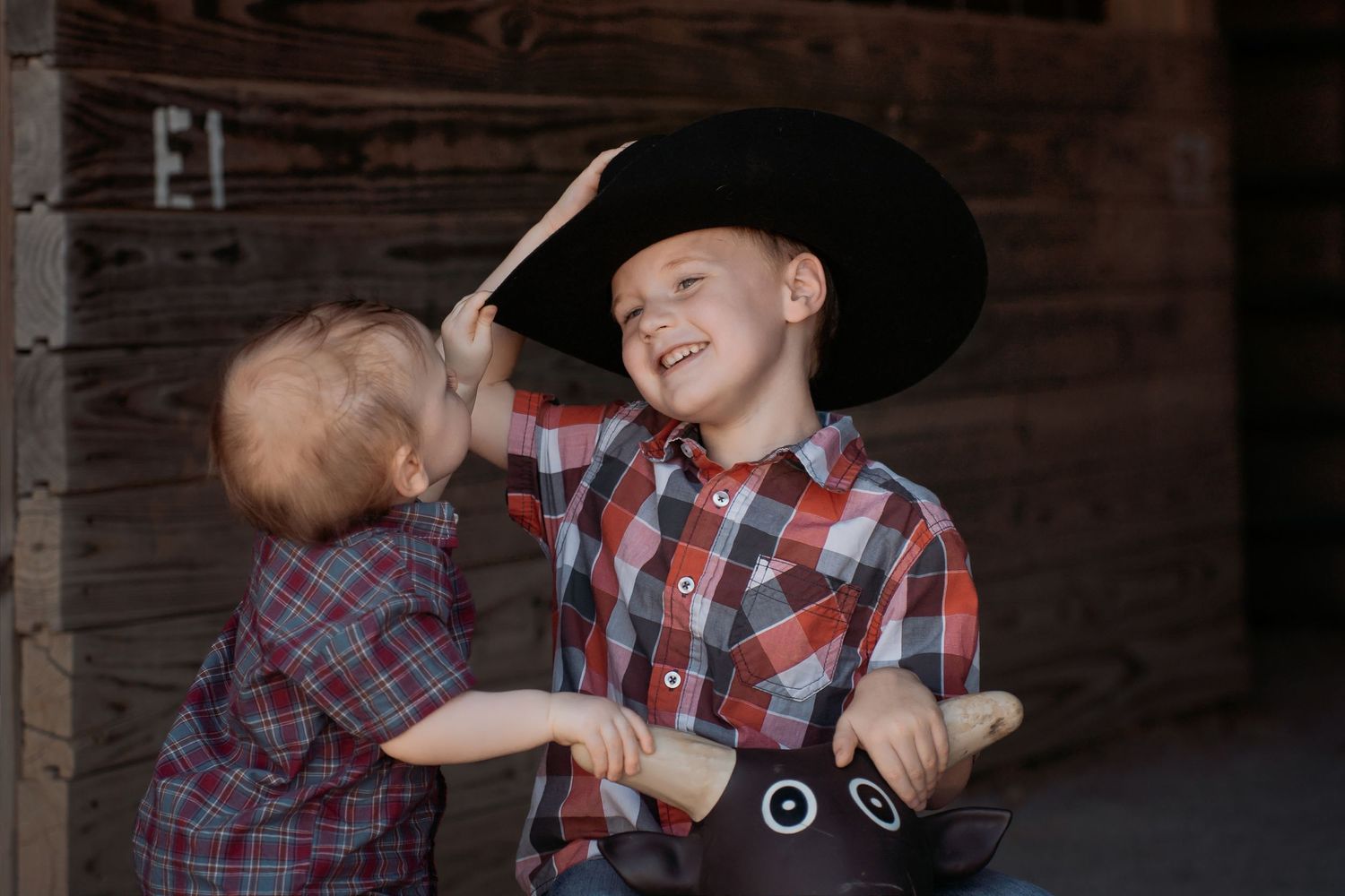 Two young boys in plaid shirts and cowboy hats play together in a rustic barn setting.