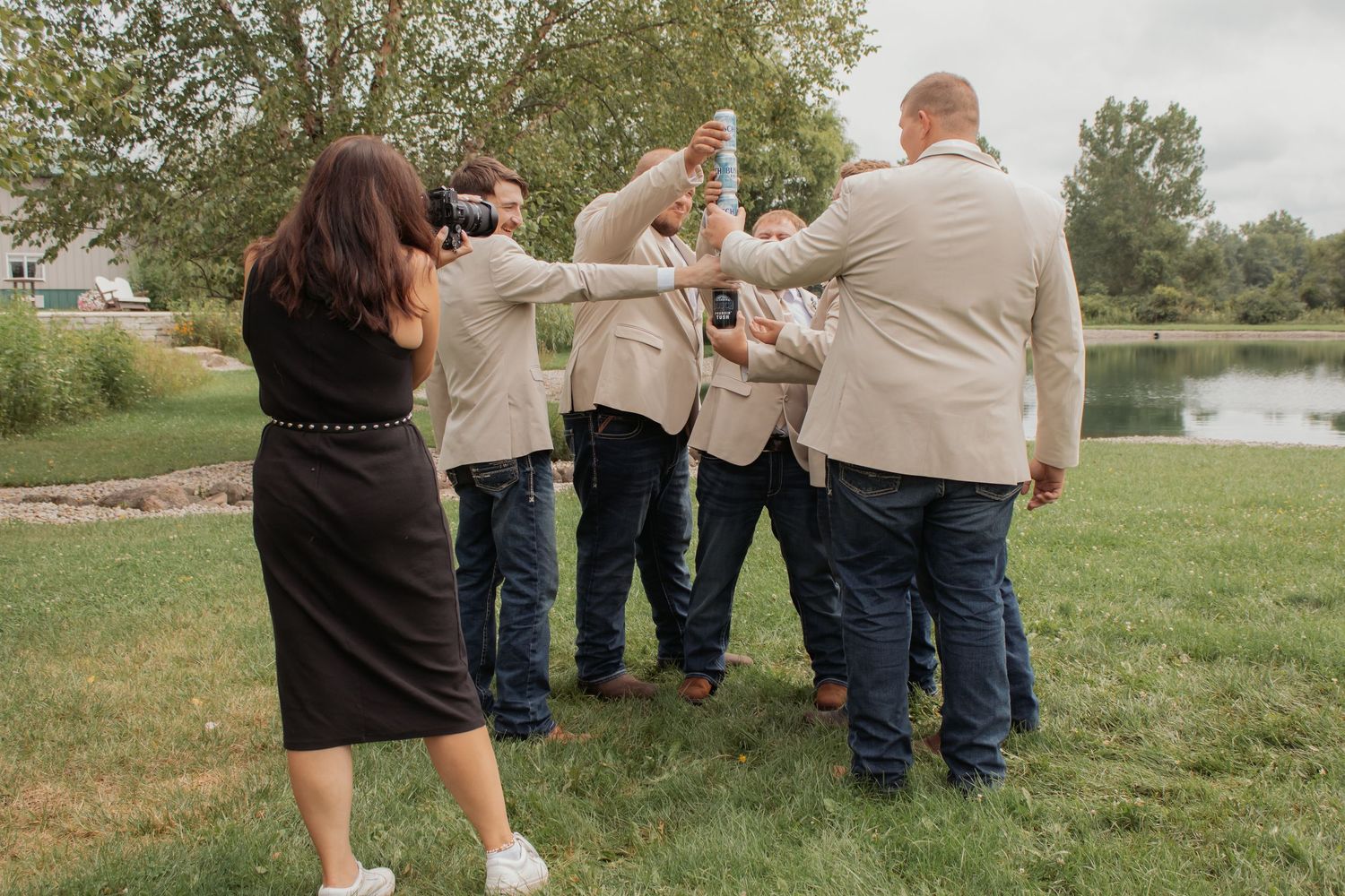 Groomsmen in tan blazers and jeans toasting drinks outdoors near a pond with a woman in a black dress.