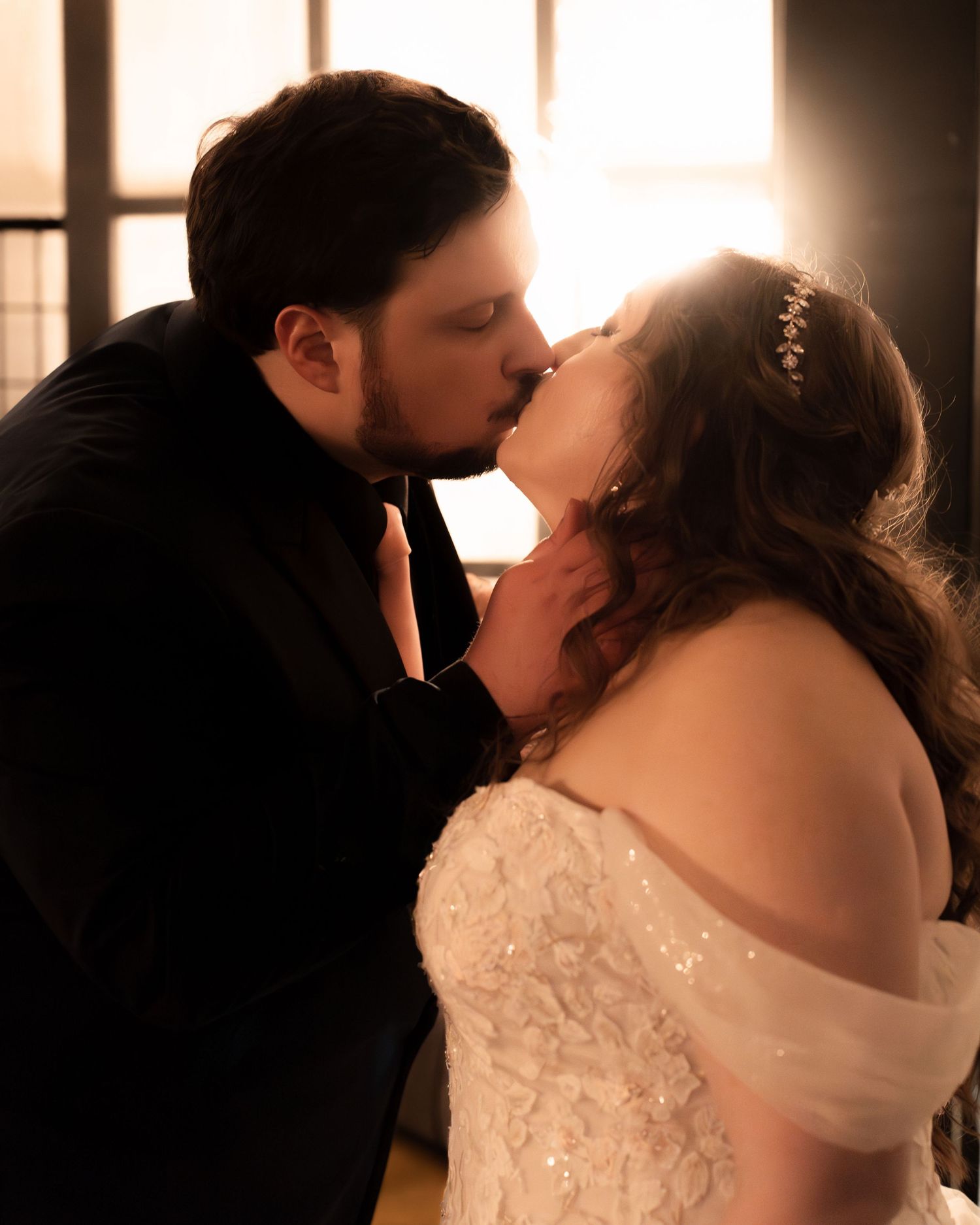 Couple sharing a romantic kiss in wedding attire, bride in lace gown with hair piece, backlit by warm golden light.