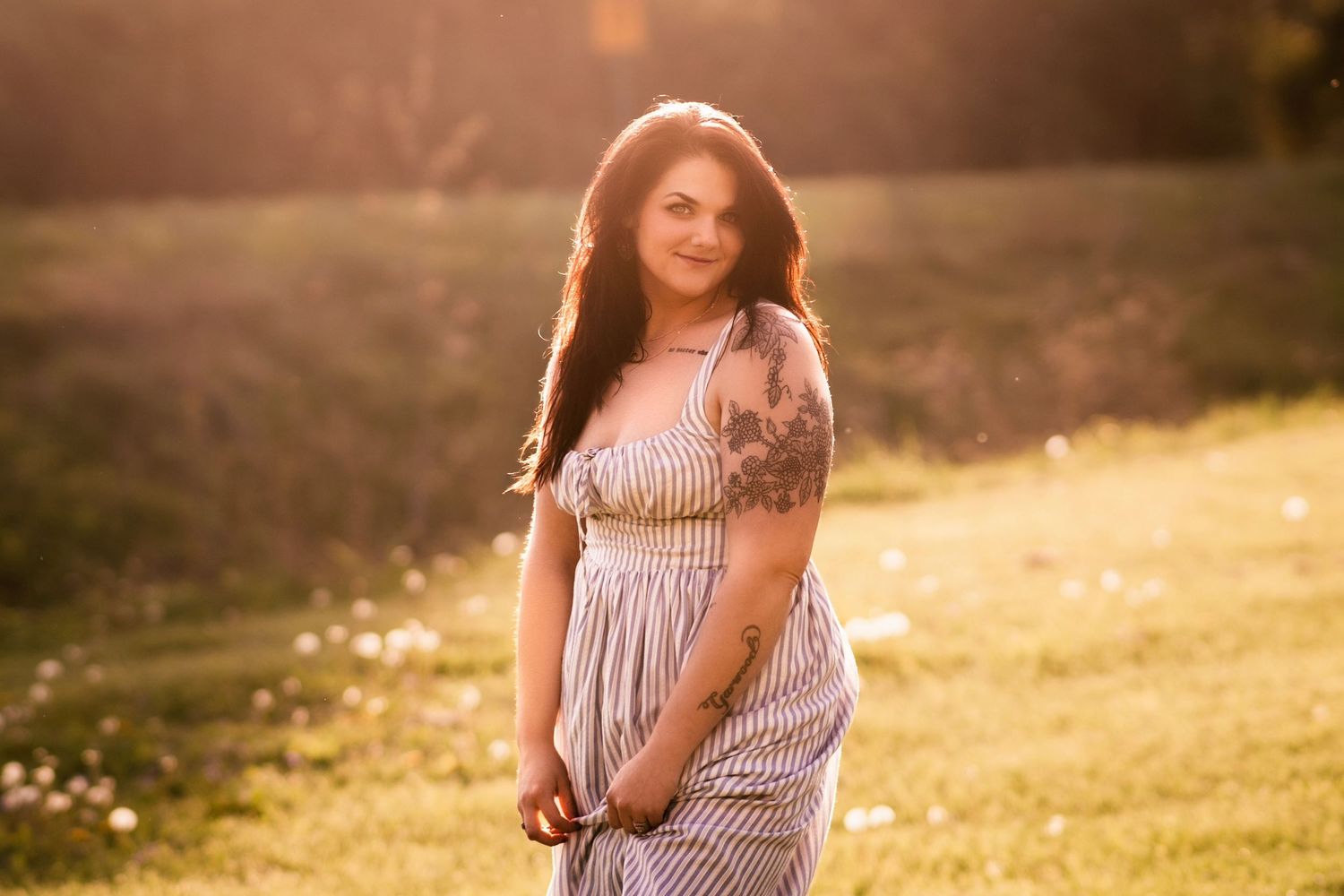 Woman in striped sundress with tattoos poses in sunlit meadow with wildflowers during golden hour.