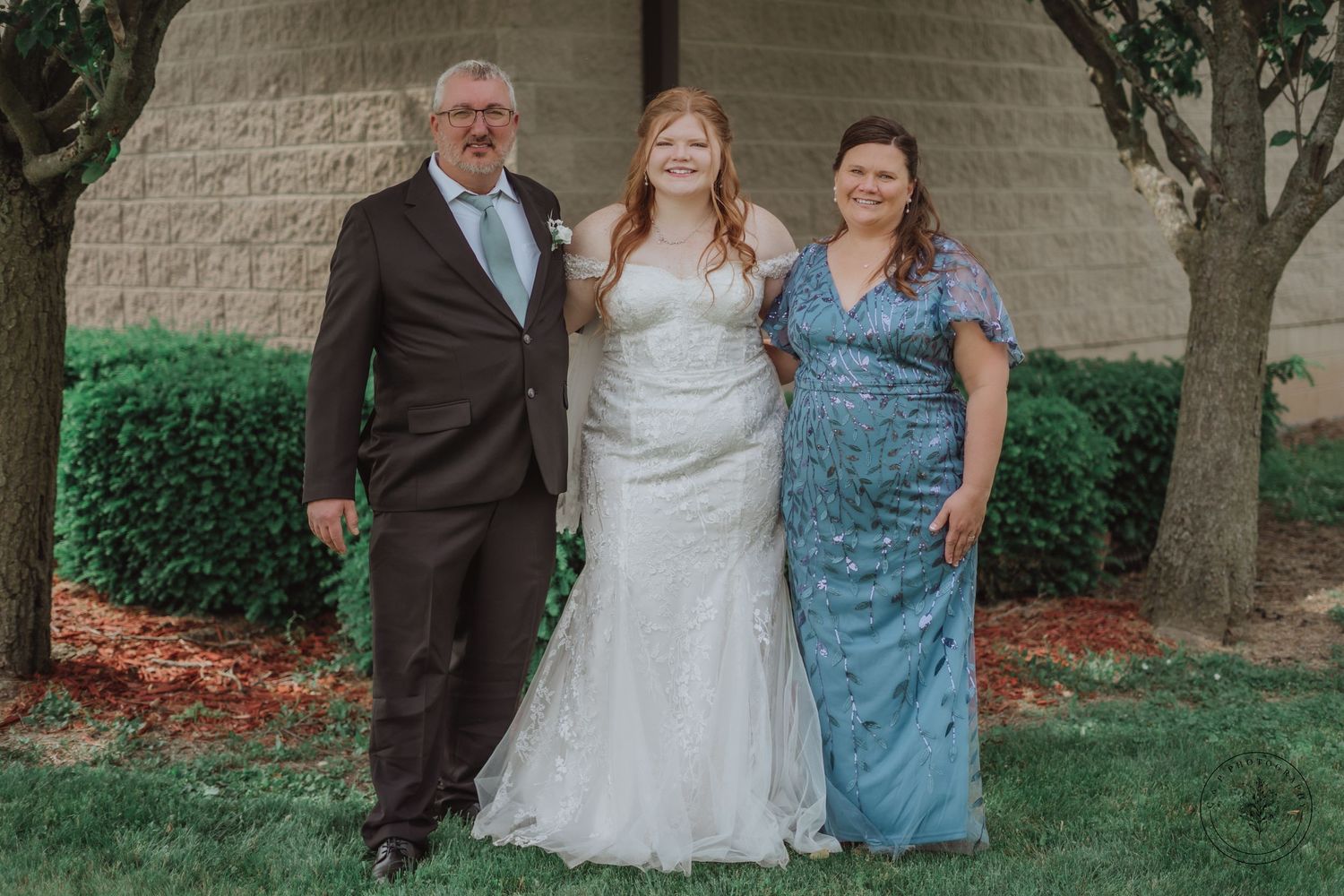 Wedding party portrait of three people in formal attire against stone wall backdrop.