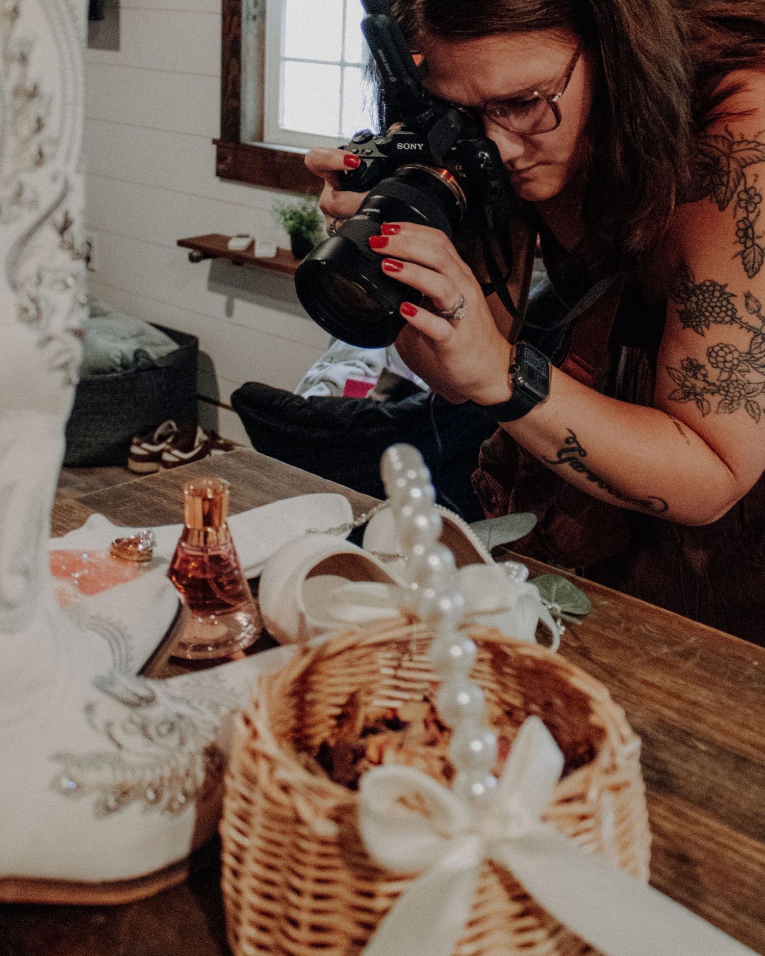 Tattooed female photographer with red nails uses Sony camera to photograph wedding details including basket and pearls on wooden table.