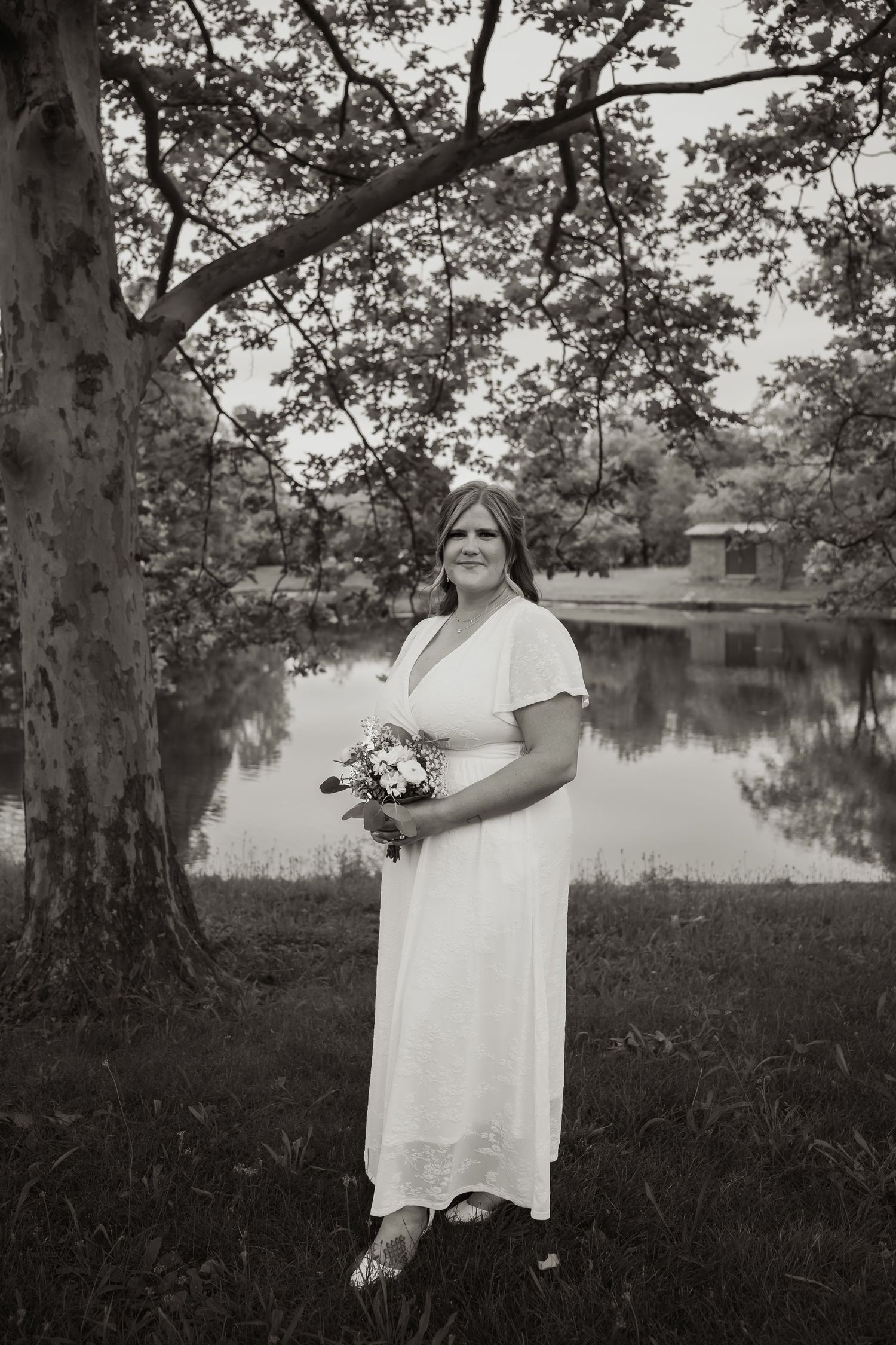Bride in white dress holding bouquet stands by pond under tree in black and white outdoor wedding portrait.