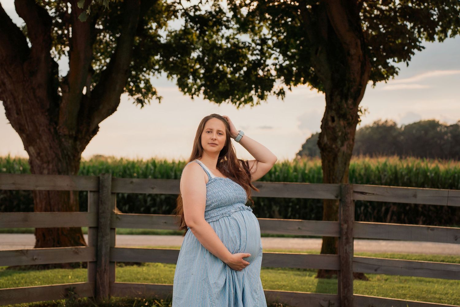 Pregnant woman in blue dress poses outdoors near wooden fence and trees during golden hour maternity photoshoot.