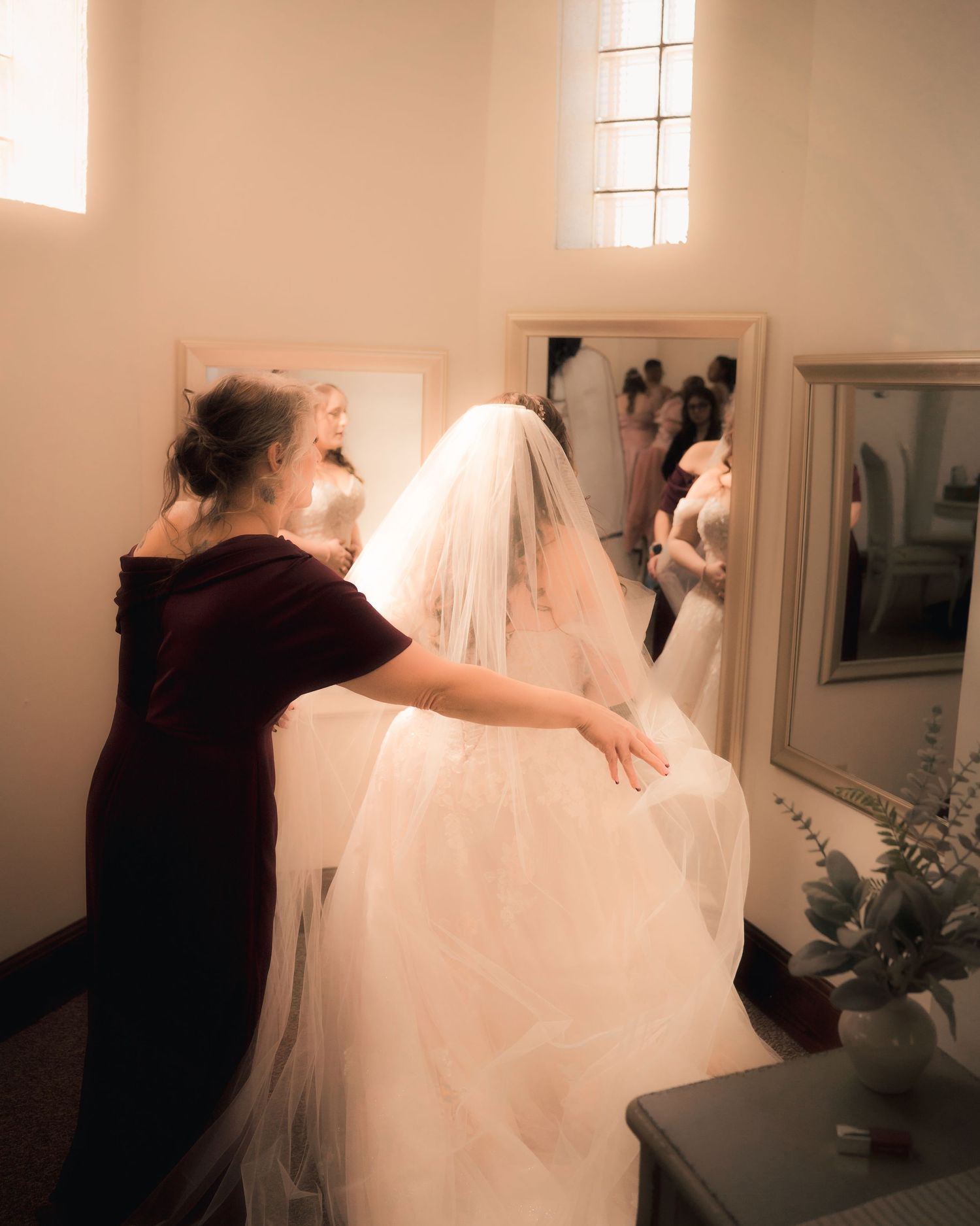 A bridesmaid in burgundy adjusts a bride's white veil and gown before the wedding ceremony, reflected in mirrors.